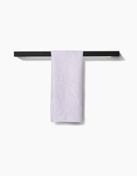 The Geometry Posh Petals Blushed Bergamot kitchen towel, in a light shade with a subtle pattern, hangs neatly over a sleek black towel bar against a white background.