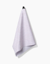 A Geometry Posh Petals Blushed Bergamot kitchen towel, featuring a light purple textured design made from recycled materials, hangs from a black hook against a plain white background.