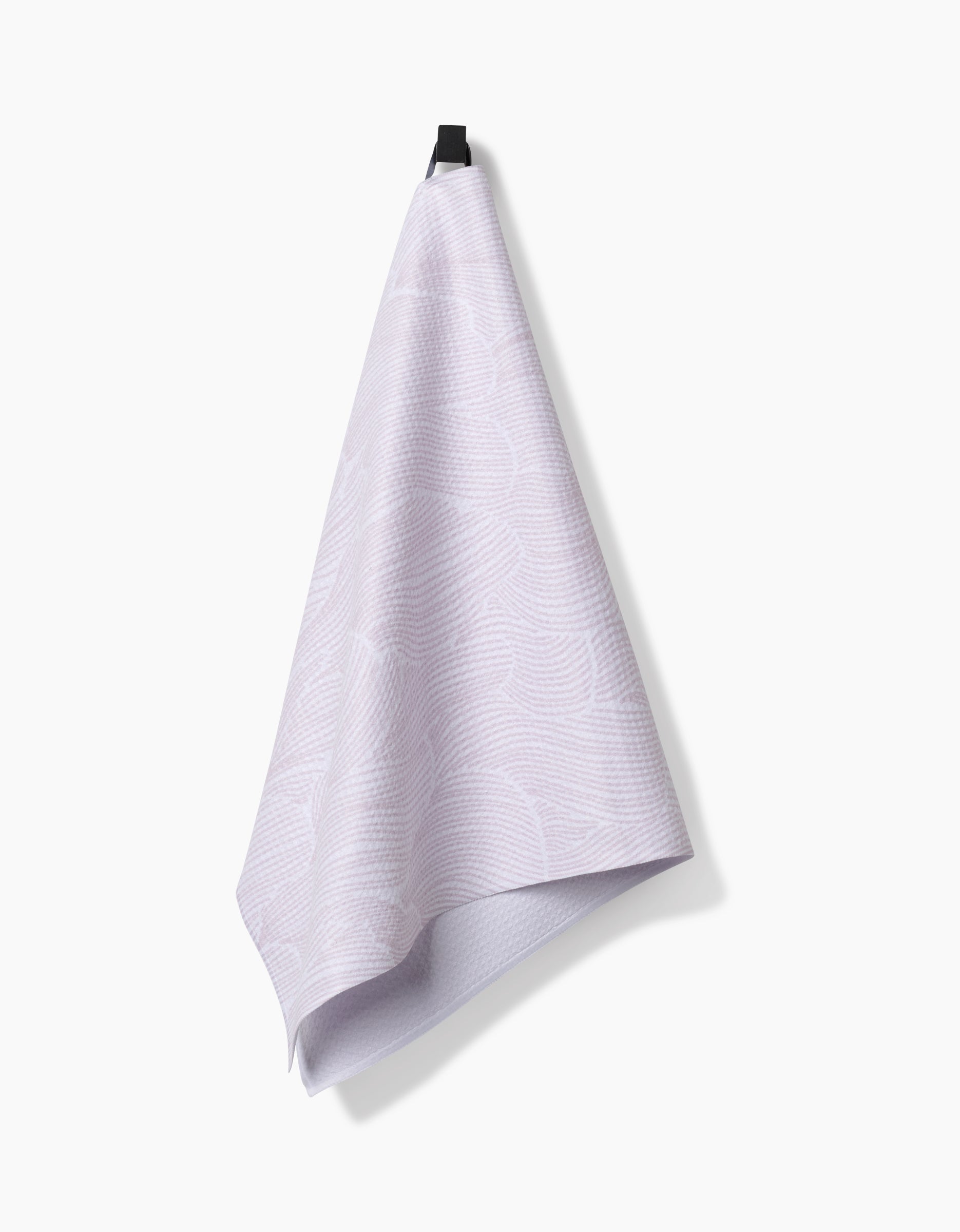 A Geometry Posh Petals Blushed Bergamot kitchen towel, featuring a light purple textured design made from recycled materials, hangs from a black hook against a plain white background.