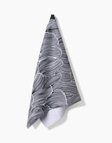 The Geometry Posh Petals Fresh Linen kitchen towel, featuring a black and white abstract wavy pattern, hangs from a black loop against a plain white background.