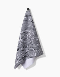 The Geometry Posh Petals Fresh Linen kitchen towel, featuring a black and white abstract wavy pattern, hangs from a black loop against a plain white background.
