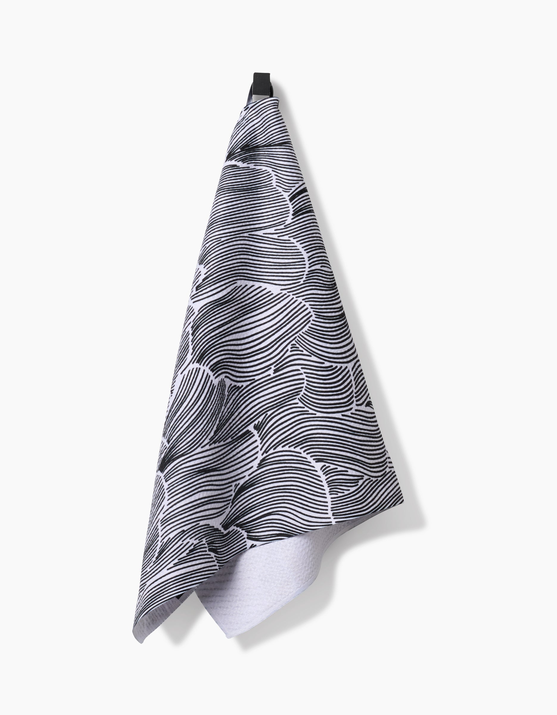 The Geometry Posh Petals Fresh Linen kitchen towel, featuring a black and white abstract wavy pattern, hangs from a black loop against a plain white background.
