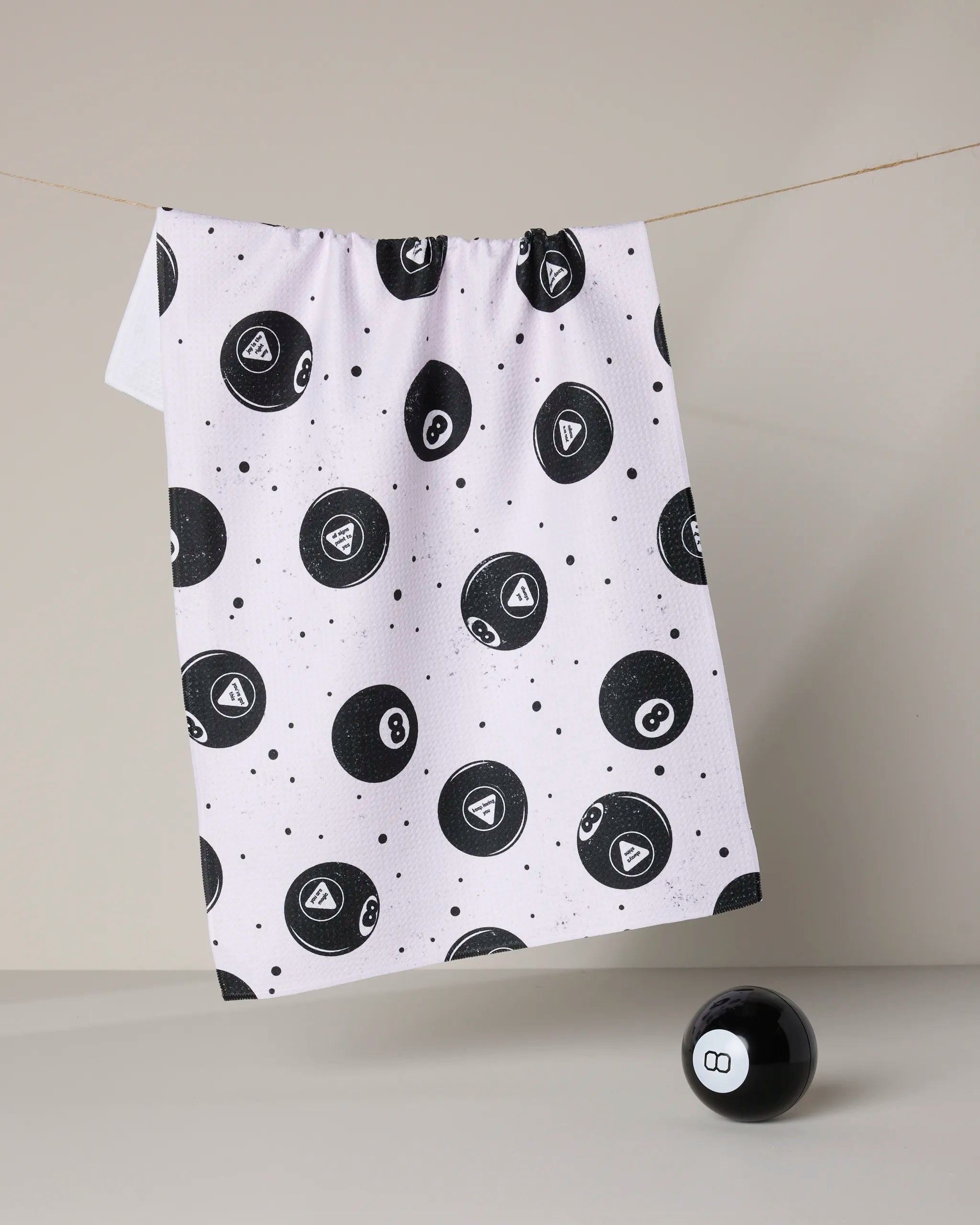 The Positive Future tea towel by Geometry, featuring a black magic 8 ball pattern, hangs on a string. Made from recycled materials, this quick-drying white dish towel is paired with a magic 8 ball toy on a neutral background.