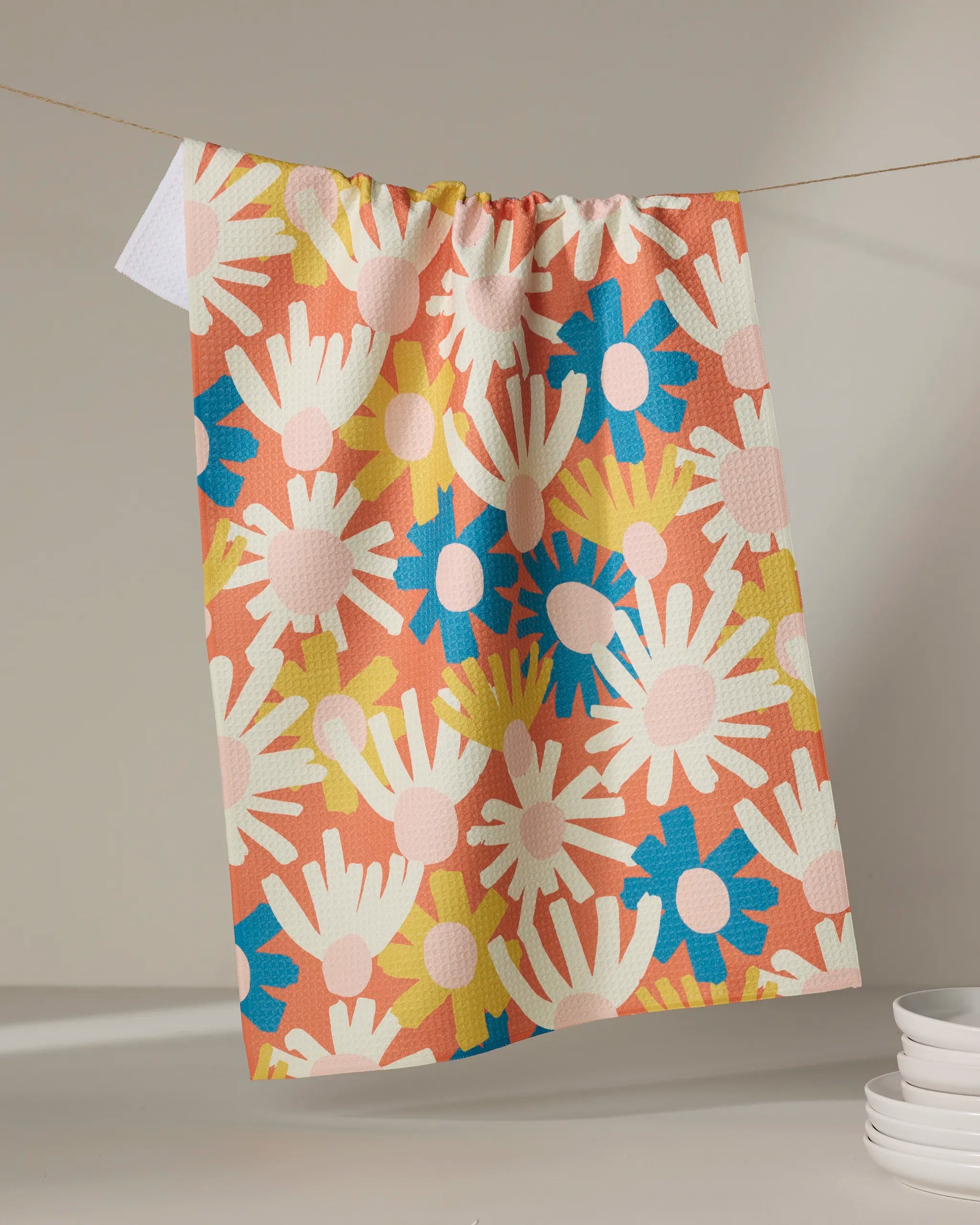 The Posy dish towel by Kelsey Weigl, featuring a colorful floral pattern on coral, hangs on a clothesline with stacked white dishes below.