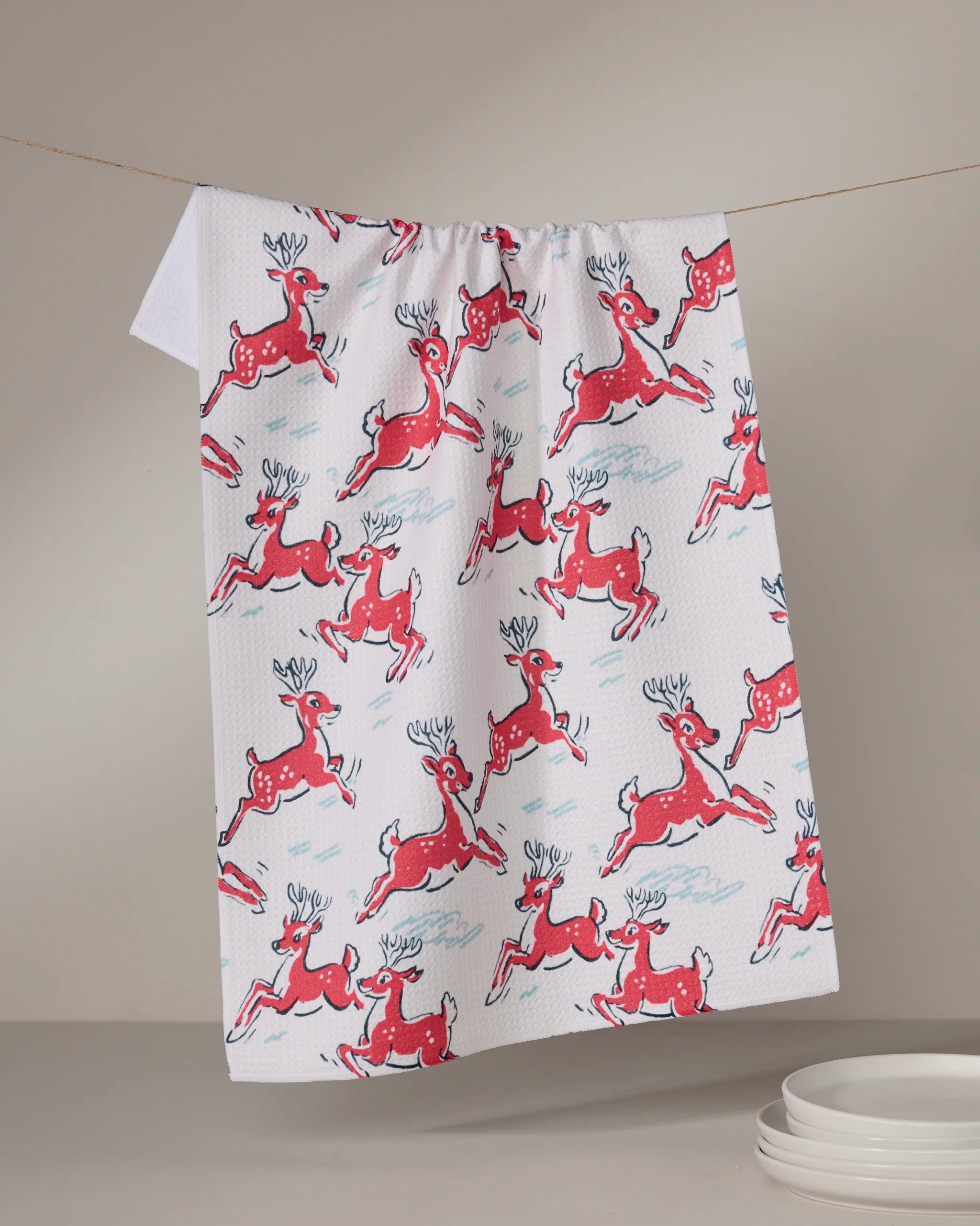 The Geometry "Prancing Reindeer" tea towel, made from Geoweave fabric and featuring red reindeer designs, hangs over a line against a neutral background with a stack of white plates in the bottom right corner.