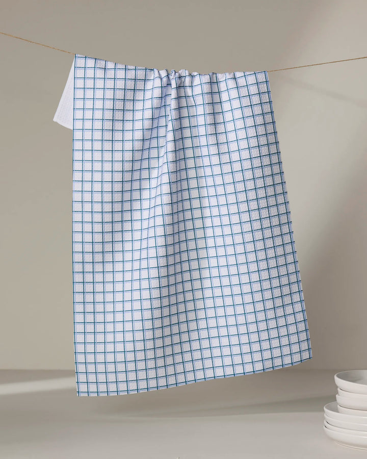 The Buy Guide’s Preppy - Emerald French Blue tea towel, made from recycled materials, hangs from a string against a neutral background with a stack of white plates partially visible in the lower right corner.