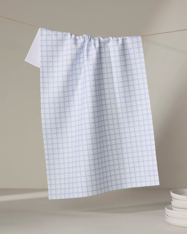 The Buy Guide's Preppy - French Blue Mint recycled kitchen towel with a light blue checkered pattern hangs on a string. A stack of white dishes is placed in the lower right corner on a light surface against a neutral background.