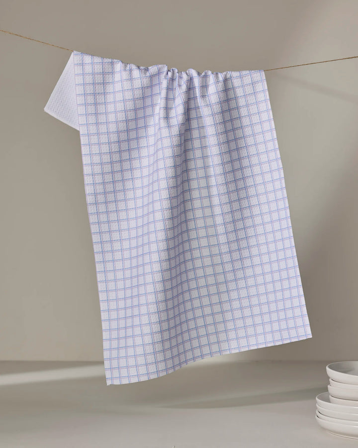 The Buy Guide's Preppy - Lilac French Blue kitchen tea towel with a blue checkered pattern hangs on a clothesline against a neutral background. A stack of white plates is partially visible. Made from recycled materials for eco-friendly style.