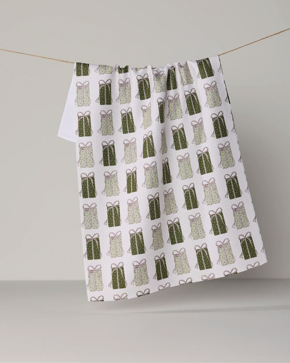 Present Parade by Geometry—a white tea towel featuring a repeating pattern of green and pink gift boxes, crafted from recycled materials—hangs neatly over a clothesline against a plain light gray background.