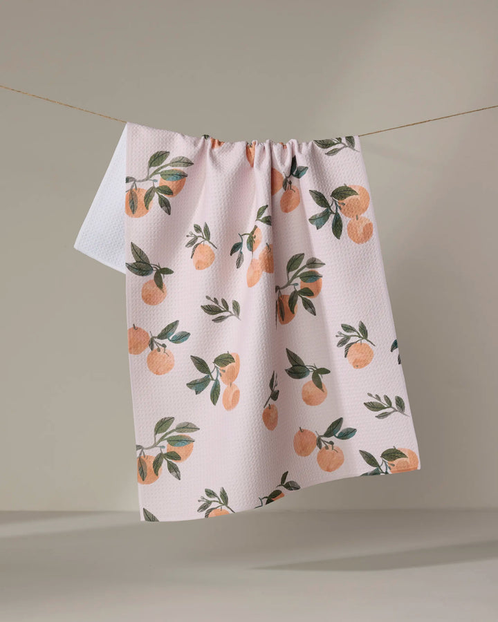 The Pretty In Peach kitchen towel by Janna Sue Design, made from recycled materials, features a peach and green leaf pattern and hangs on a clothesline against a neutral background.