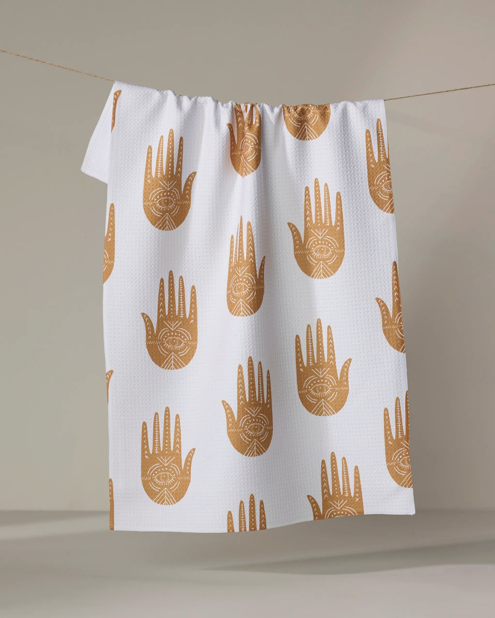 The "Protection" tea towel by Jessica Swift is a white, quick-drying towel with a repeating gold-brown hamsa hand and eye design, shown hanging on a clothesline against a plain background.