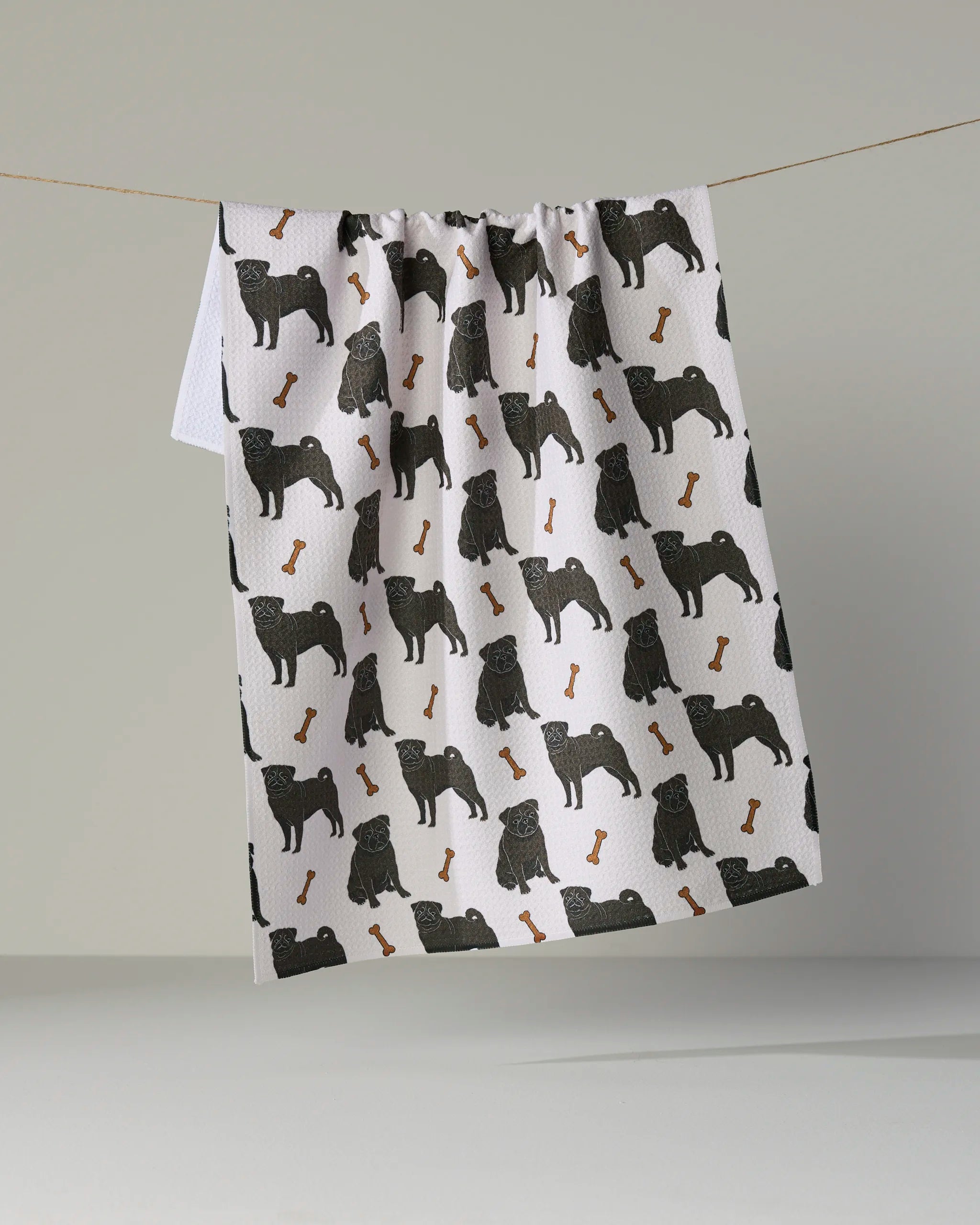 Here is a revised version of your sentence using the provided product data: The Pug Club tea towel by Geometry features a playful repeating pattern of black pug silhouettes and orange bones on a white background, hanging on a string against a plain, light gray backdrop. Made from recycled materials, this eco-friendly kitchen towel brings charming style and sustainability to your home.