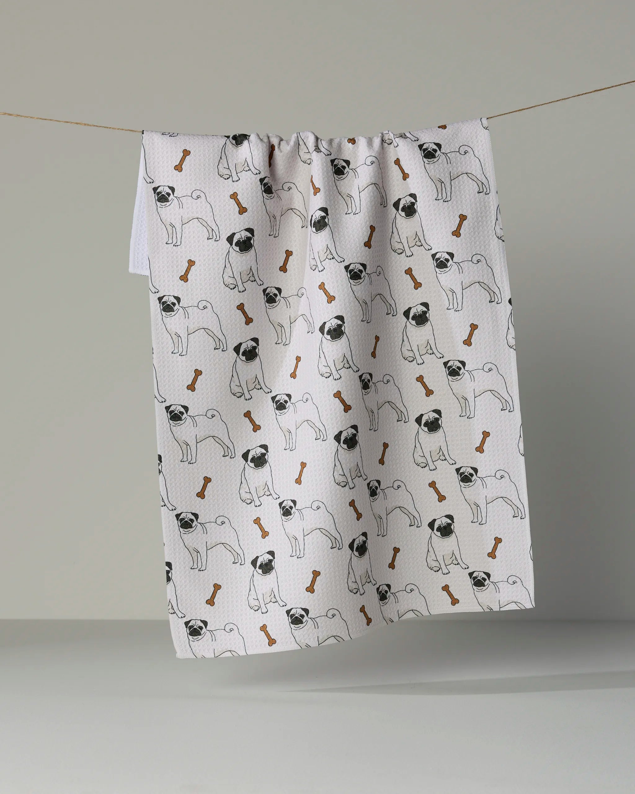 A “Pug Club” kitchen towel by Geometry, featuring an adorable pattern of pugs and brown dog bones on a white background, hangs on a string against a plain gray backdrop. This charming tea towel brings both style and practicality to any kitchen.