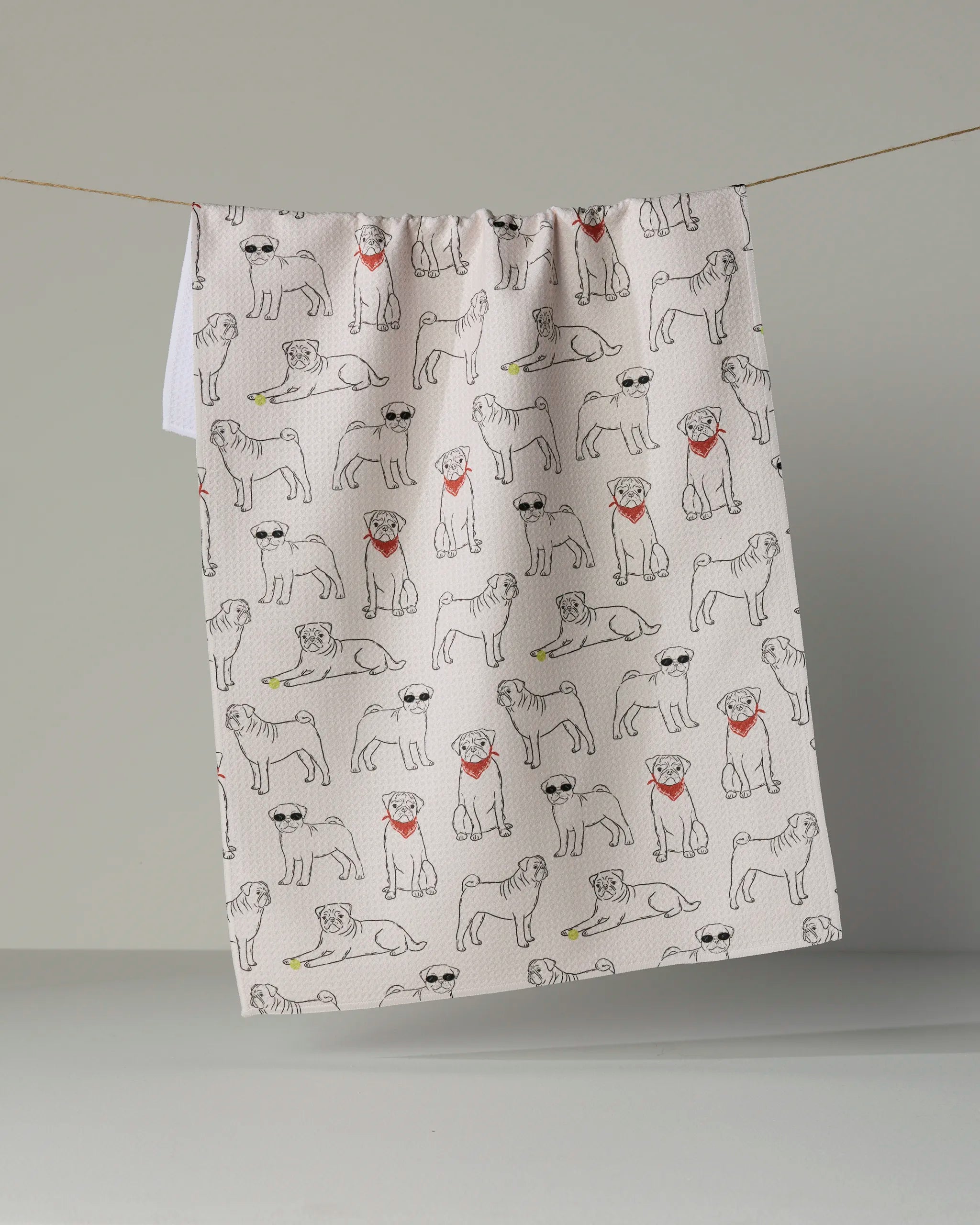 A white recycled kitchen towel from Geometry, called “Pug Pals,” hangs on a line, featuring a playful pattern of illustrated pugs—some sporting red scarves or sunglasses, and others holding green bones in their mouths.