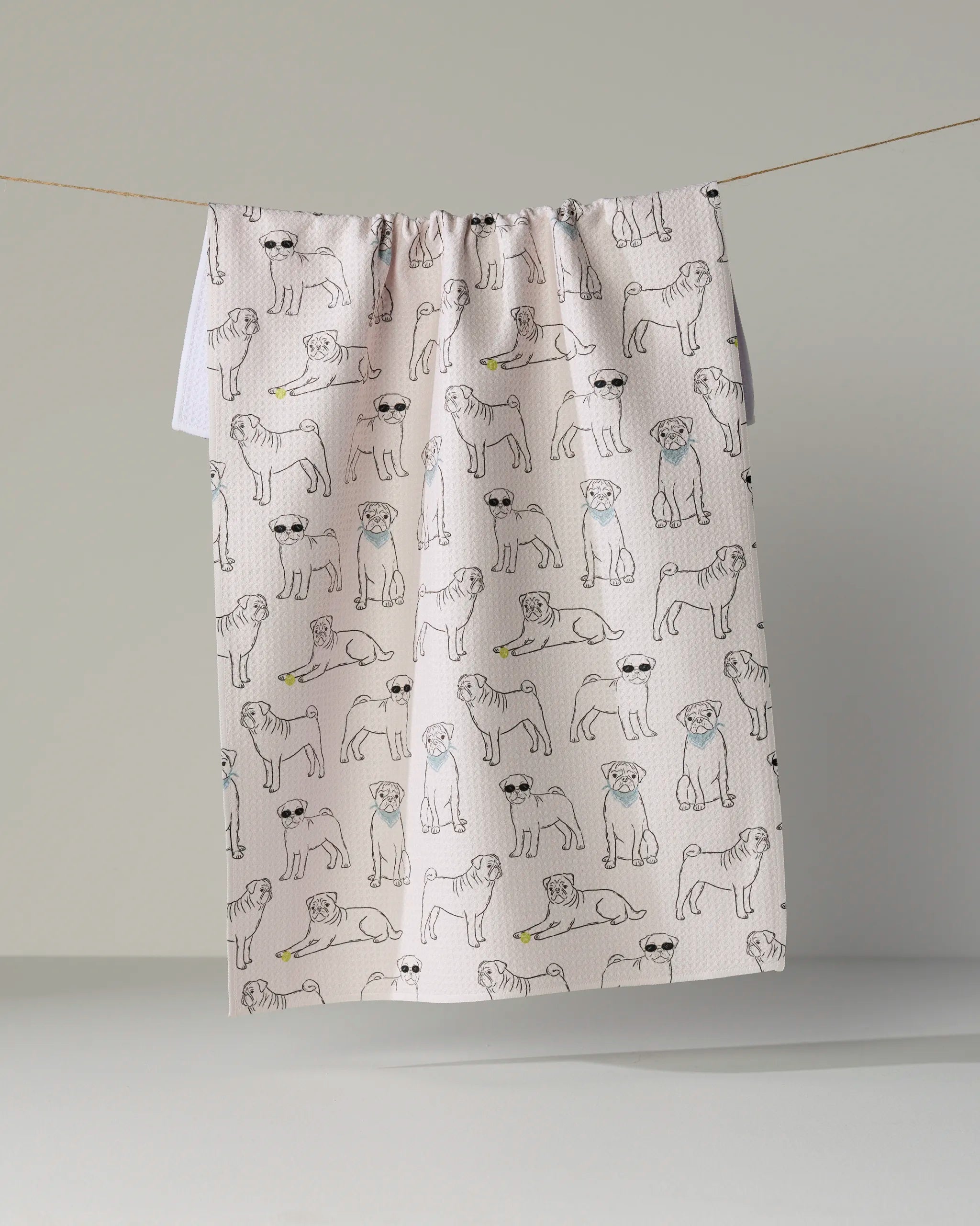 The Geometry “Pug Pals” tea towel is a white, ultra-absorbent towel adorned with line-drawn pugs wearing sunglasses and chewing yellow bones, shown hanging from a string against a simple light background.