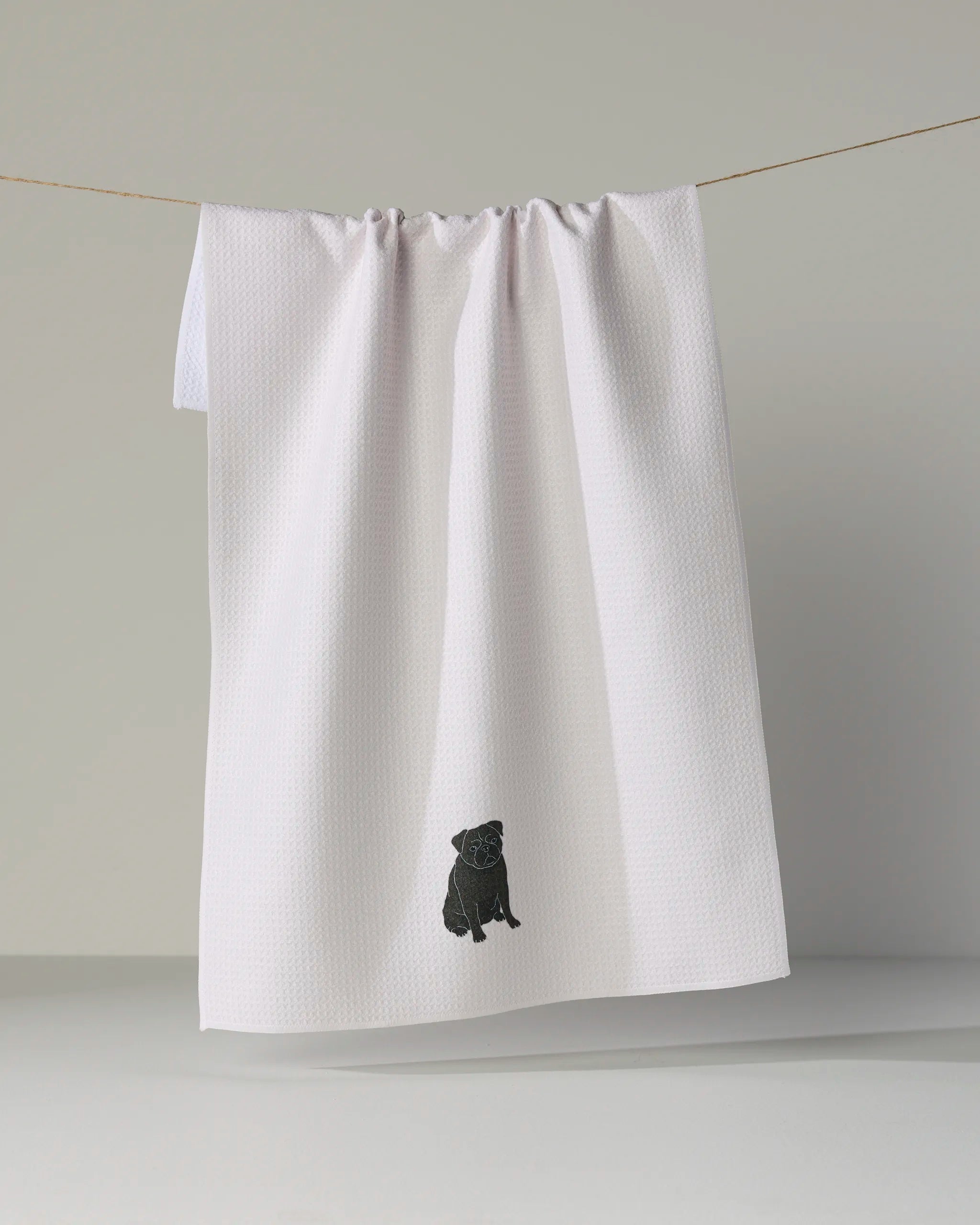 A white textured tea towel from Geometry, called “Pug Pose,” hangs on a line, featuring a small black embroidered dog near the bottom center. Crafted from recycled materials, this ultra-absorbent towel combines eco-friendly function with charming design.