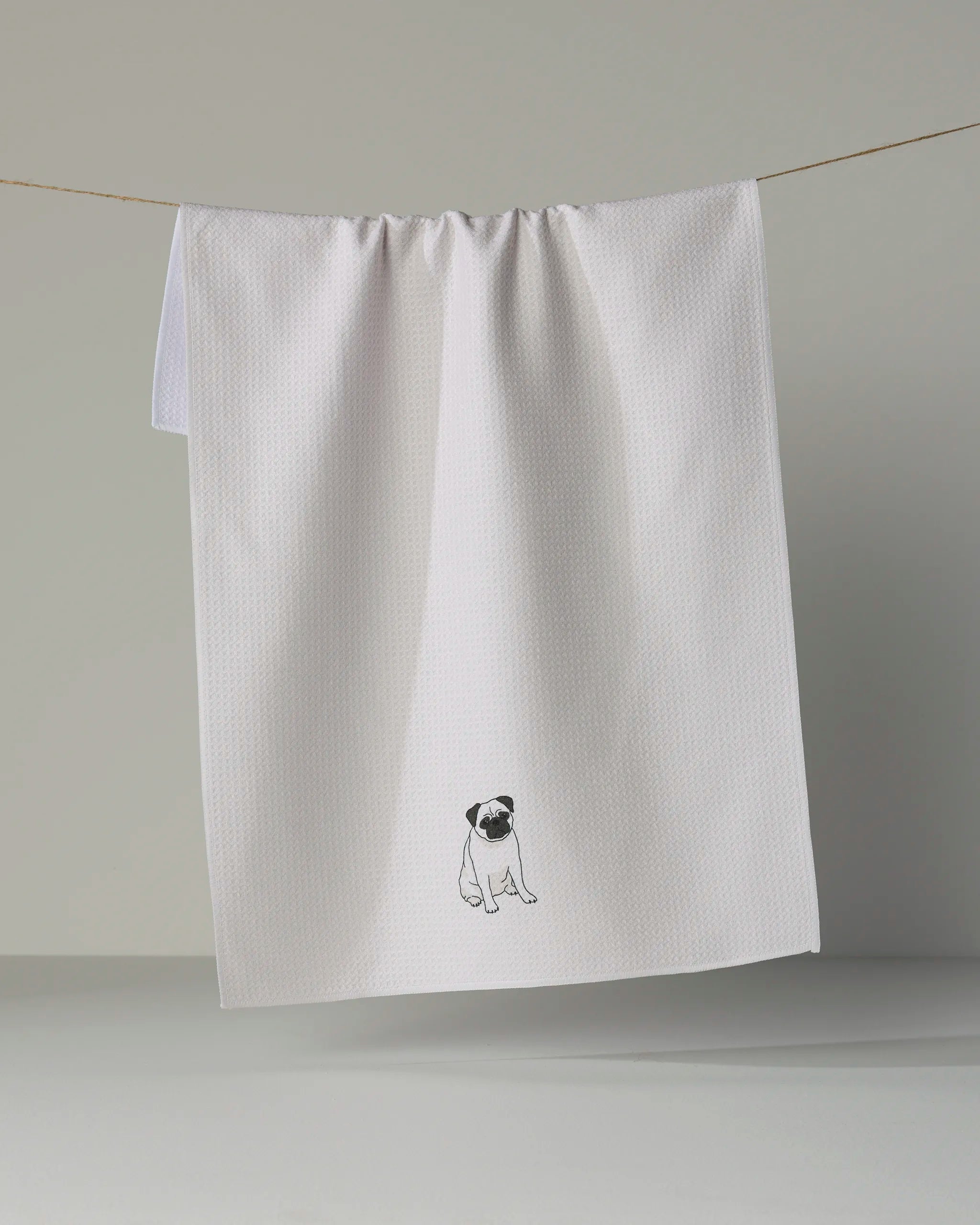 A white textured tea towel by Geometry, called “Pug Pose,” hangs on a line and features a small embroidered pug near the bottom center, crafted from ultra-absorbent recycled materials.