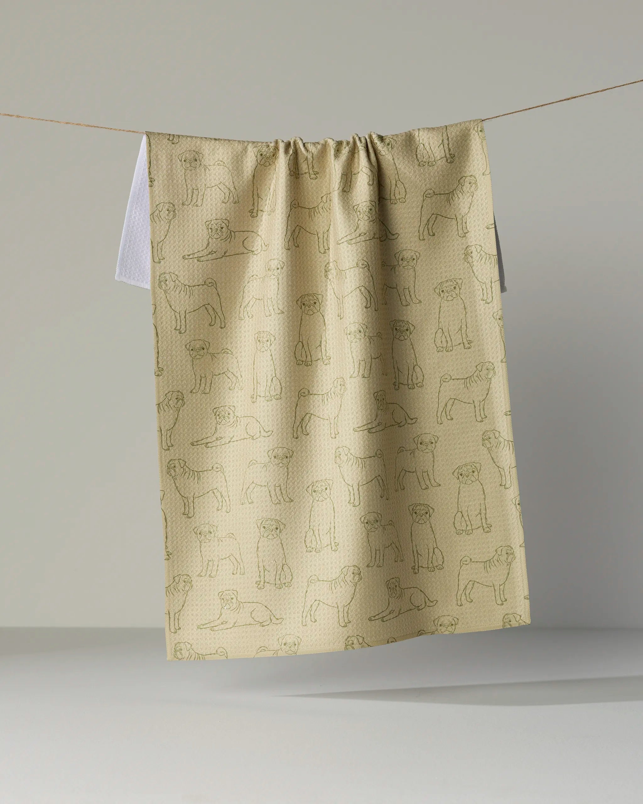 A “Pug Pups” kitchen tea towel by Geometry, featuring an outline pattern of pugs in beige and made from recycled materials, hangs on a clothesline against a plain background.