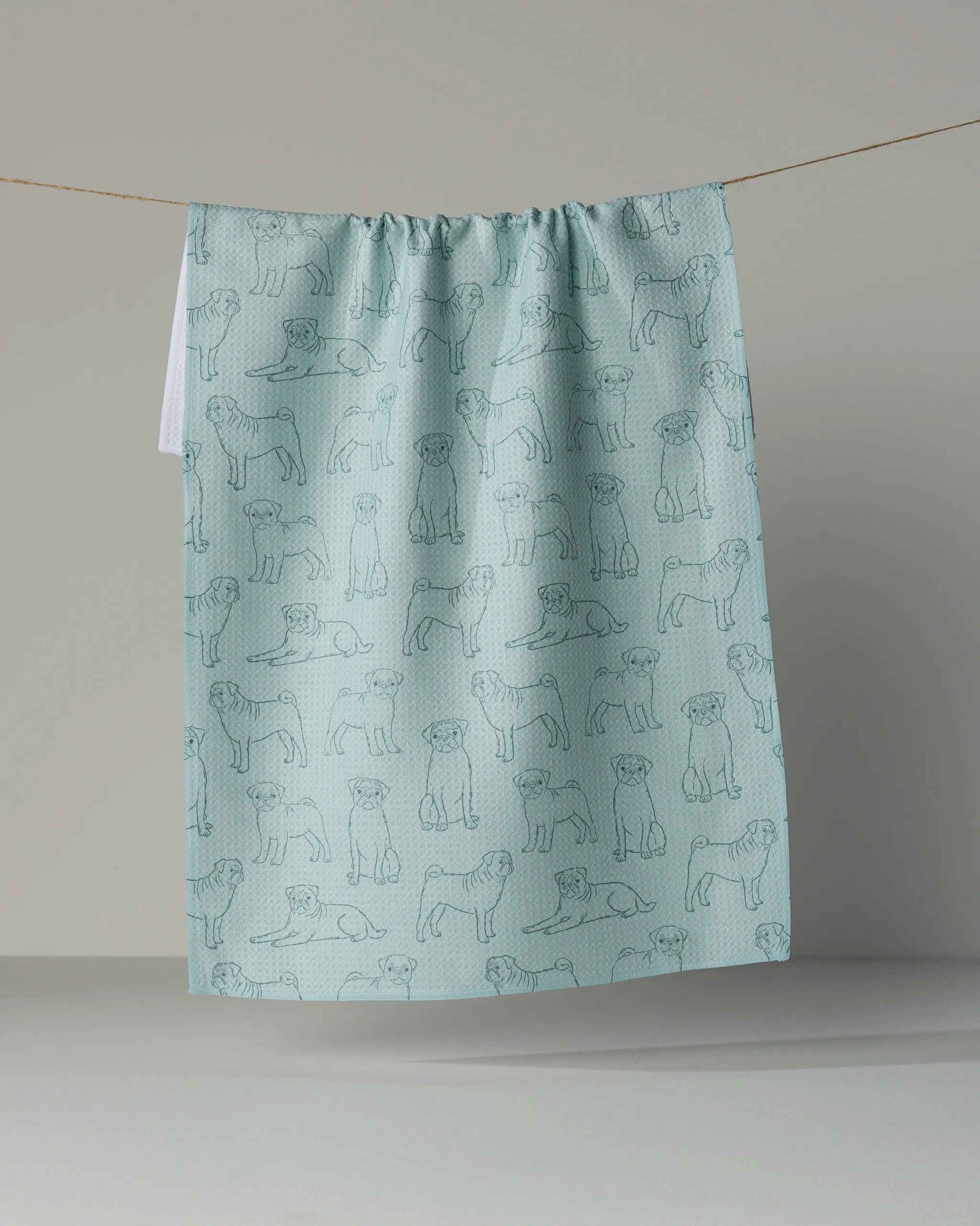 The “Pug Pups” kitchen tea towel by Geometry, crafted from recycled materials, showcases an outline pattern of playful pugs in different poses on a light blue background and is displayed hanging on a clothesline against a plain backdrop.