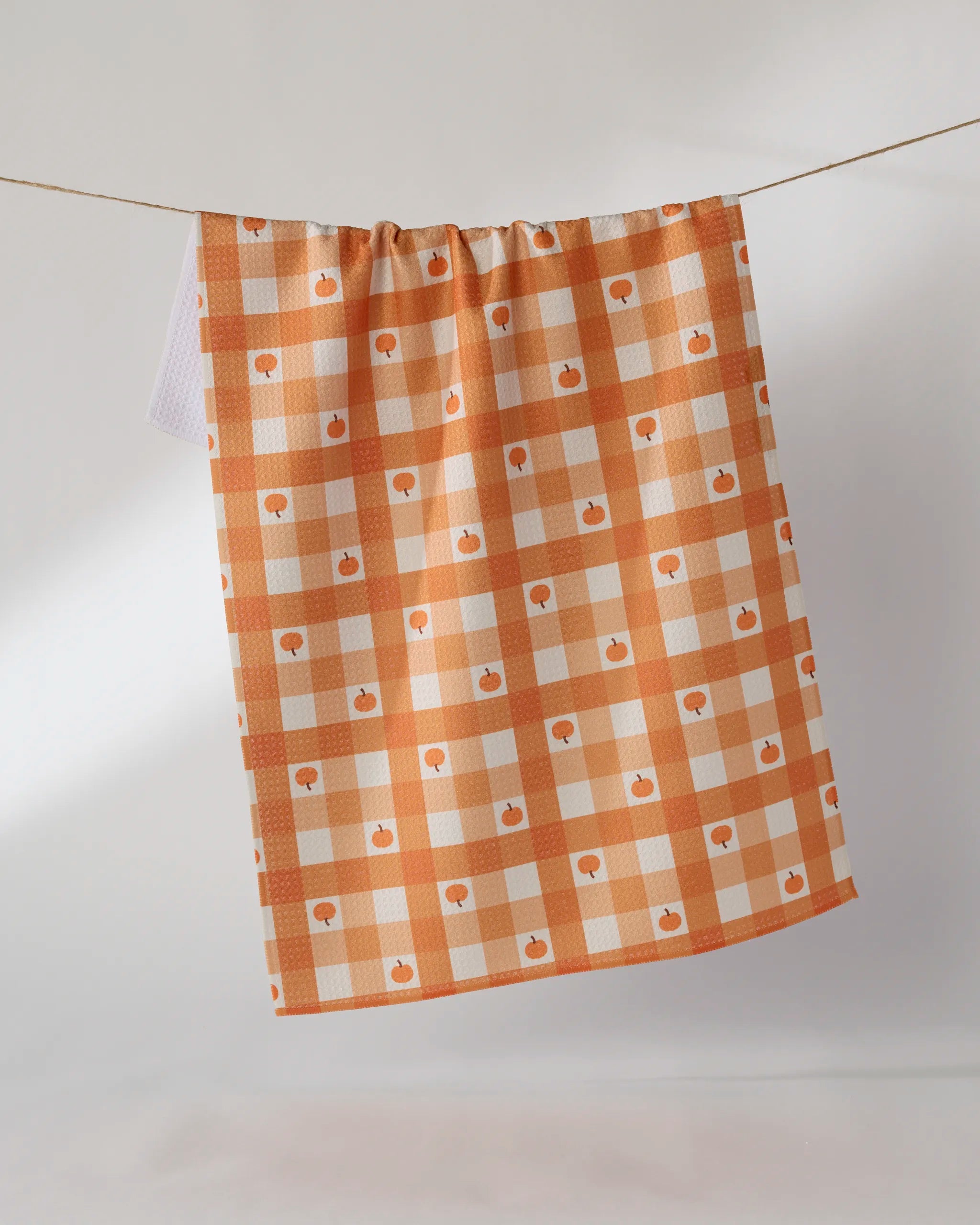 The Geometry Pumpkin Check tea towel, made from recycled materials, features an orange and white checkered pattern with small apple icons and hangs on a clothesline against a plain, light background.