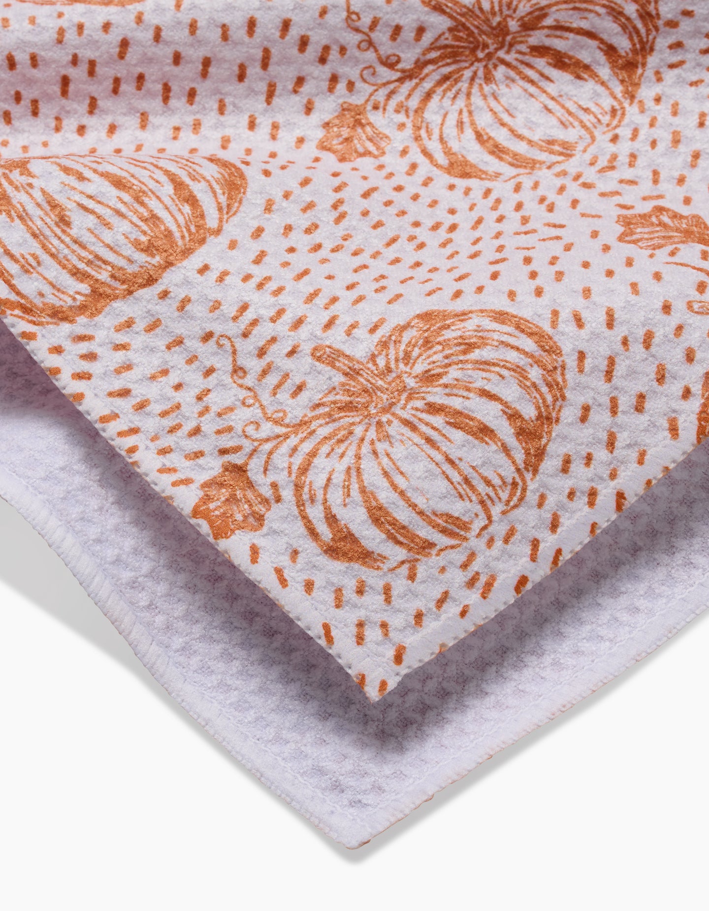 A close-up of the Pumpkin Field kitchen towel by Charlie Rowan Designs, featuring a white background with orange pumpkins and dots, partially folded to highlight its soft, textured fabric made from recycled materials.