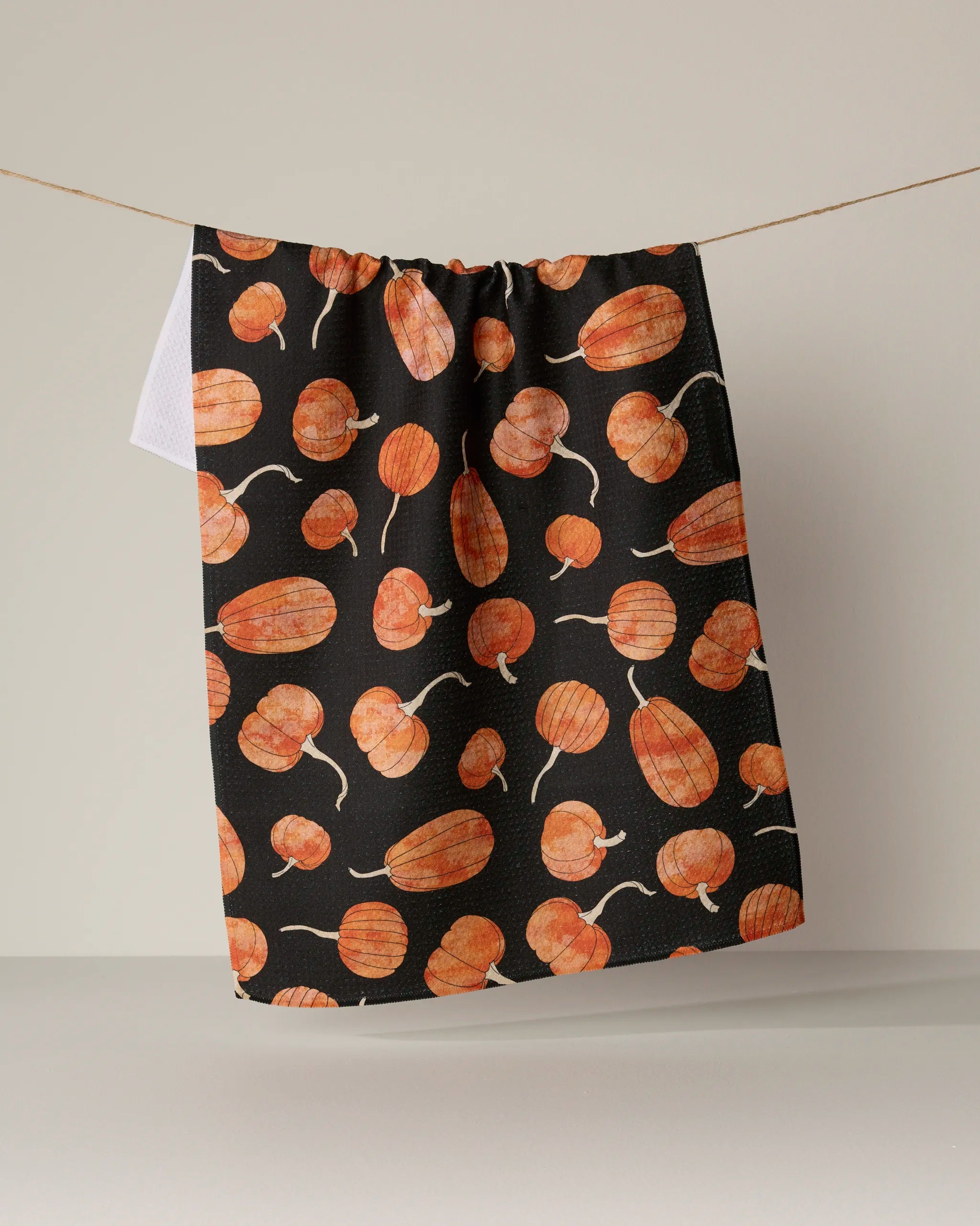 A Geometry “Pumpkin Hollow” ultra-absorbent kitchen tea towel, featuring an orange pumpkin pattern on a black background, hangs on a clothesline against a plain, neutral wall.