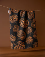 The Geometry Pumpkin Noir recycled kitchen towel, featuring an orange pumpkin pattern on black, hangs from a string against a brown background.