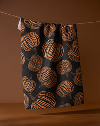 The Geometry Pumpkin Noir recycled kitchen towel, featuring an orange pumpkin pattern on black, hangs from a string against a brown background.