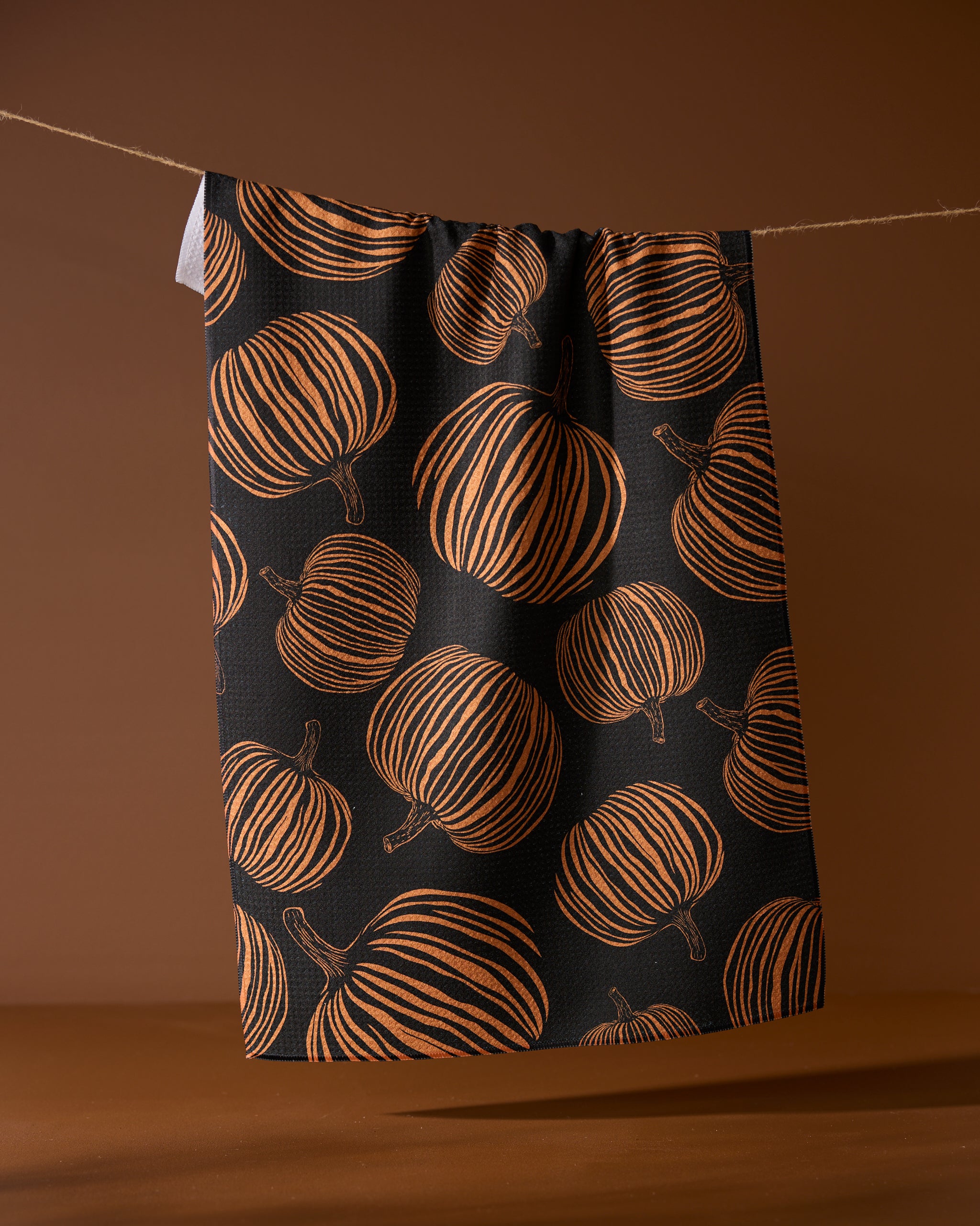 The Geometry Pumpkin Noir recycled kitchen towel, featuring an orange pumpkin pattern on black, hangs from a string against a brown background.