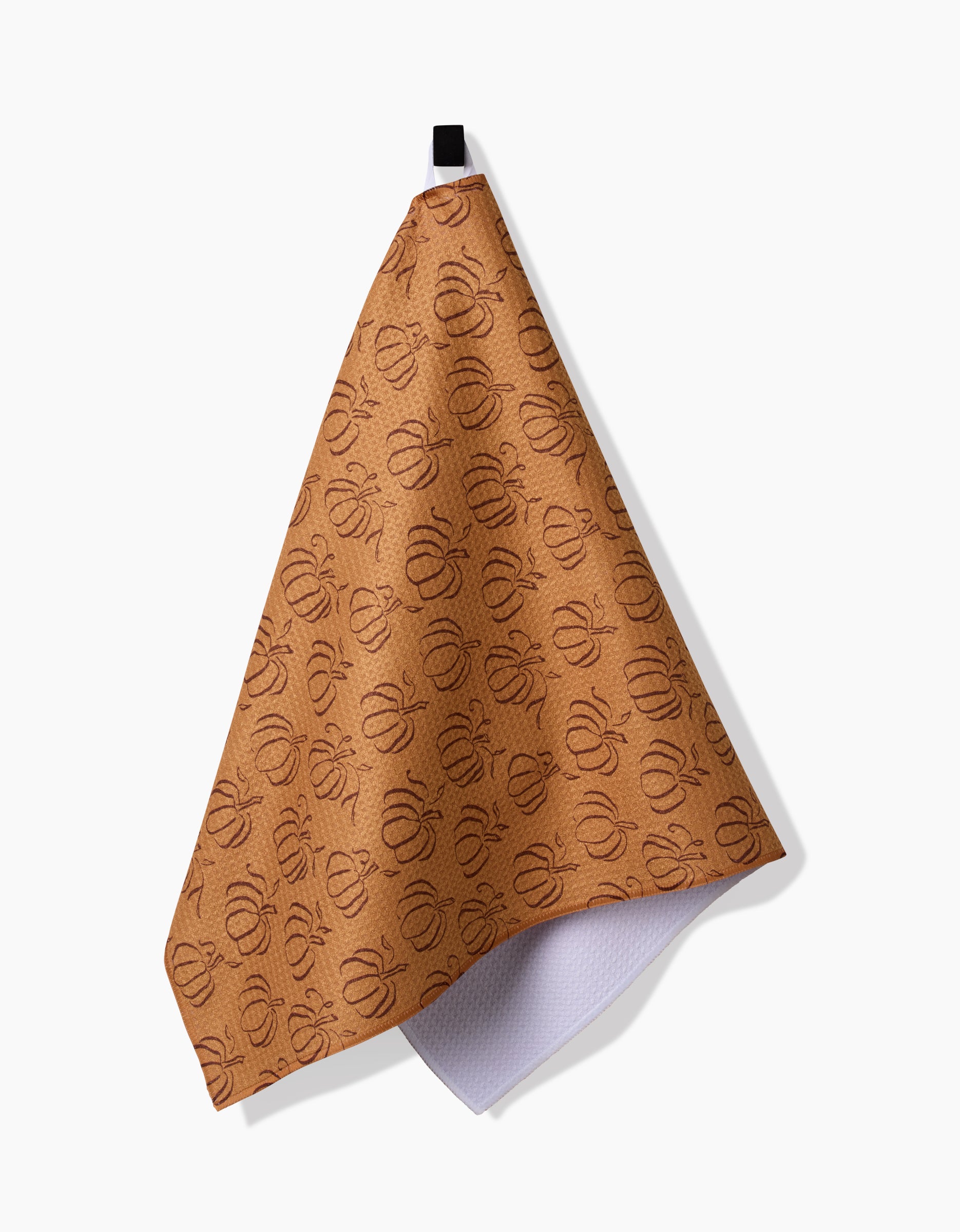 Aubrey Fairchild’s Pumpkin Patchwork kitchen towel hangs from a black loop against a white background, revealing its white underside at the corner. The towel features a brown recycled fabric with a repeating patchwork pattern.