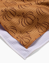 Close-up of Aubrey Fairchild’s Pumpkin Patchwork brown tea towel featuring a pumpkin pattern, layered over a white towel; both display visible edge stitching.