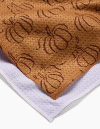 Close-up of Aubrey Fairchild’s Pumpkin Patchwork brown tea towel featuring a pumpkin pattern, layered over a white towel; both display visible edge stitching.
