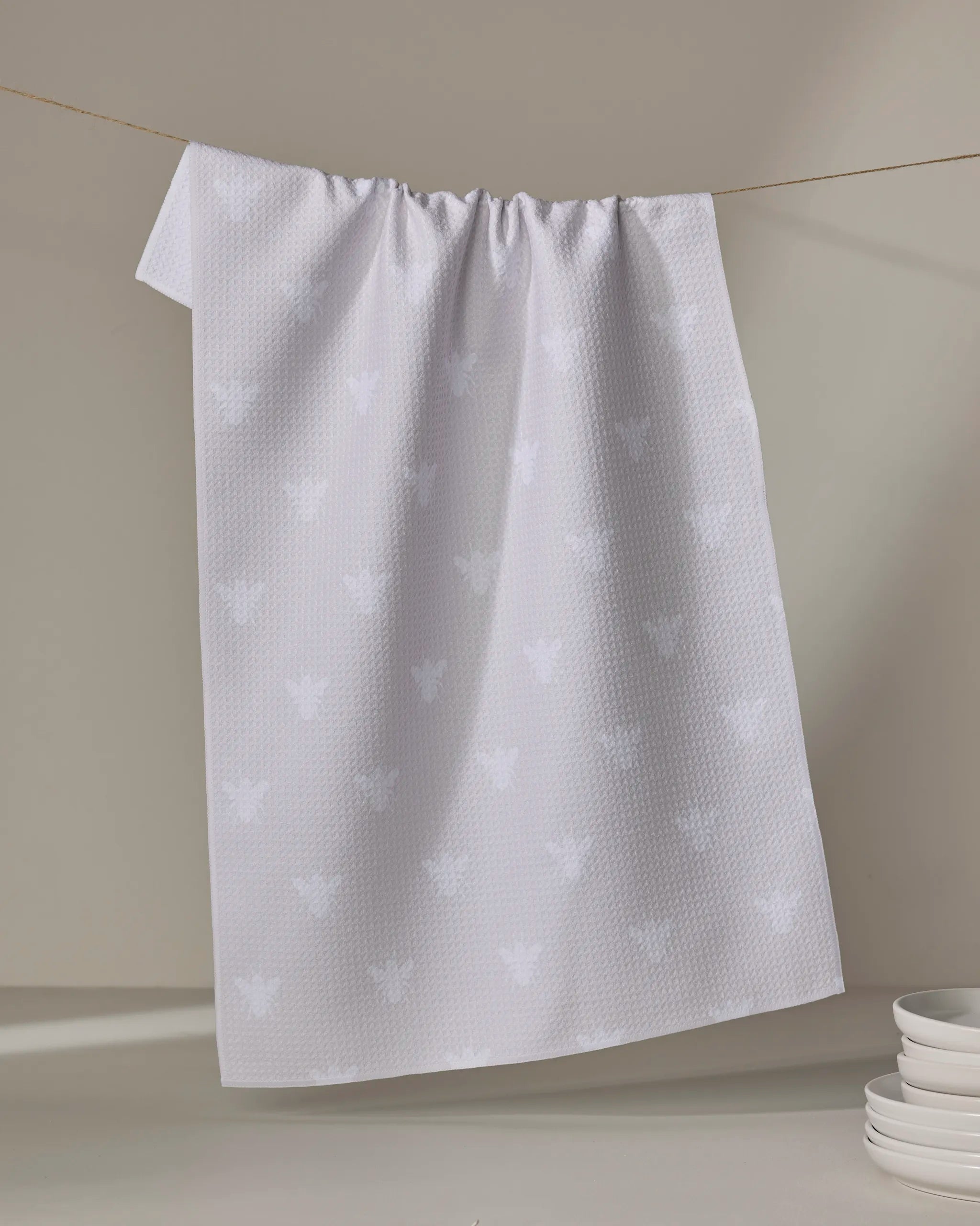 The Buy Guide’s “Queen Bee - Beige” kitchen towel, crafted from recycled materials and featuring a subtle bee pattern, hangs against a neutral background with stacked white plates in the corner.