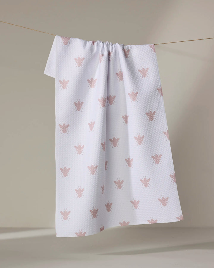 The Buy Guide’s Queen Bee - Dusty Rose blanket, made from recycled materials, features a white textured design with light pink bees and is shown hanging on a clothesline against a neutral backdrop.