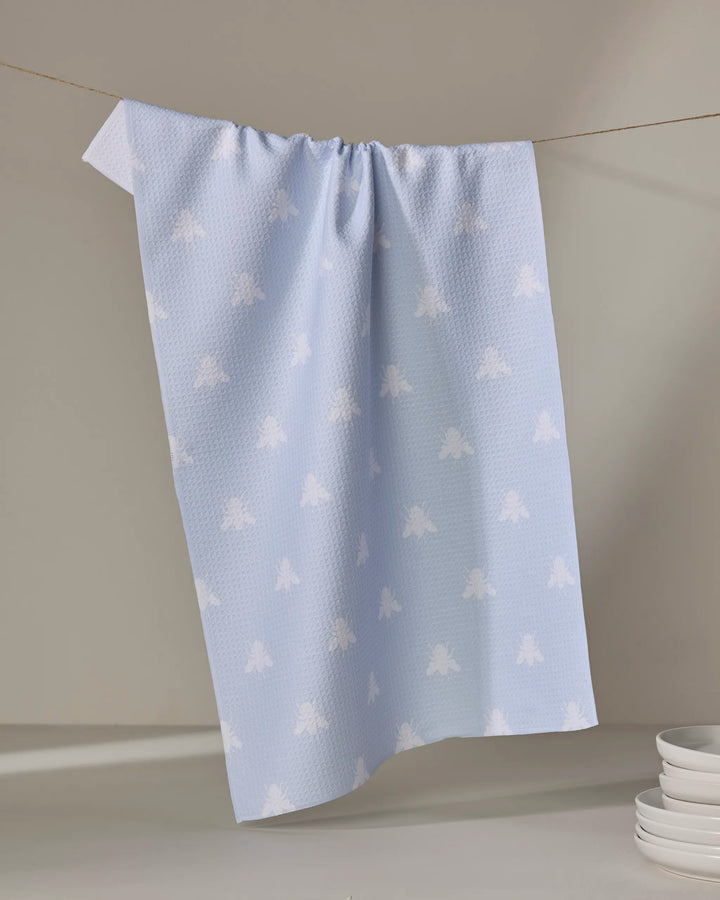 The Buy Guide’s Queen Bee - Light Blue kitchen tea towel, made from recycled materials, features a subtle white bee pattern and hangs on a string near stacked white plates against a neutral backdrop.