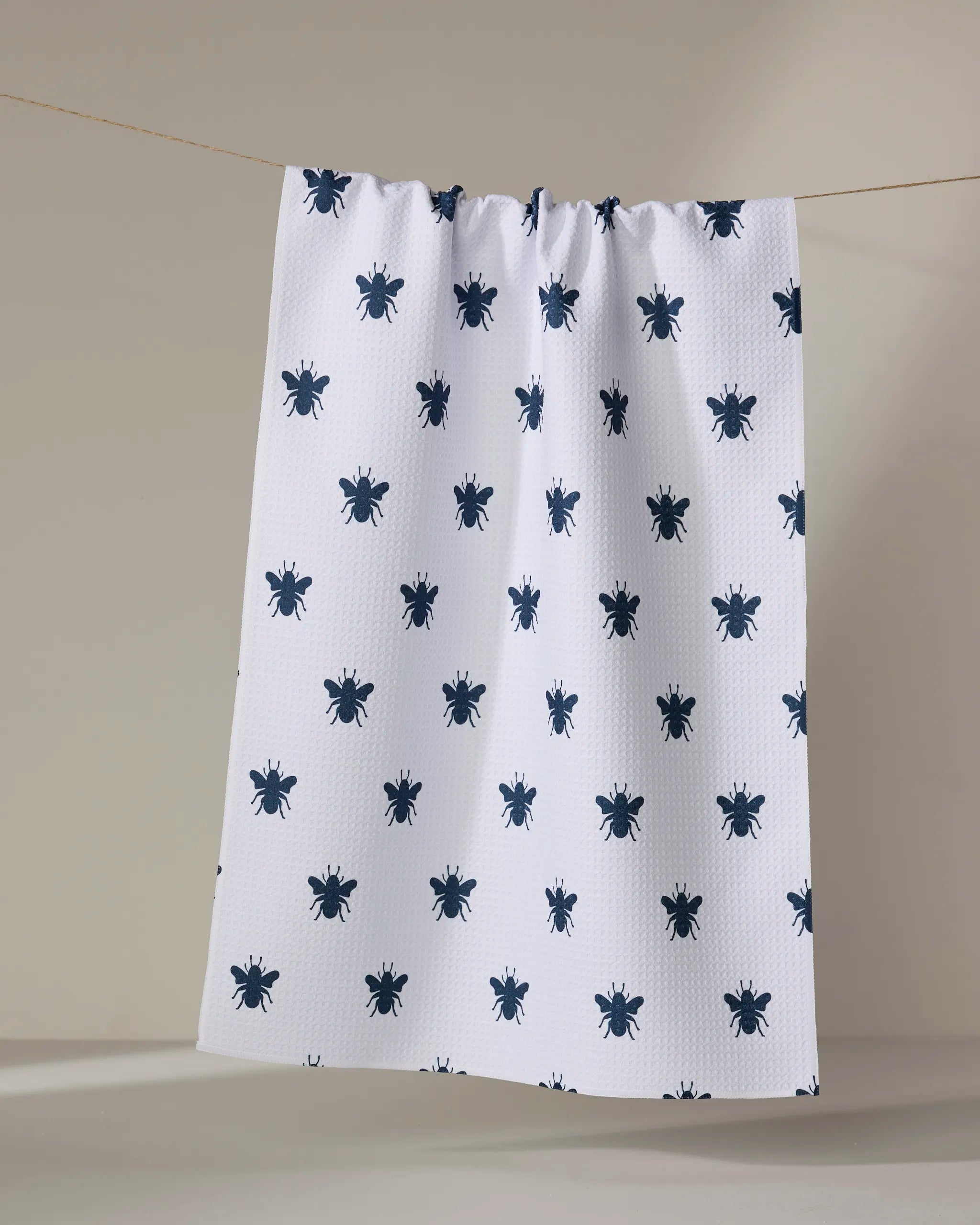 The Buy Guide's "Queen Bee - Navy" tea towel, featuring a repeating pattern of dark blue bee illustrations on white fabric, hangs from a string against a plain, neutral background.