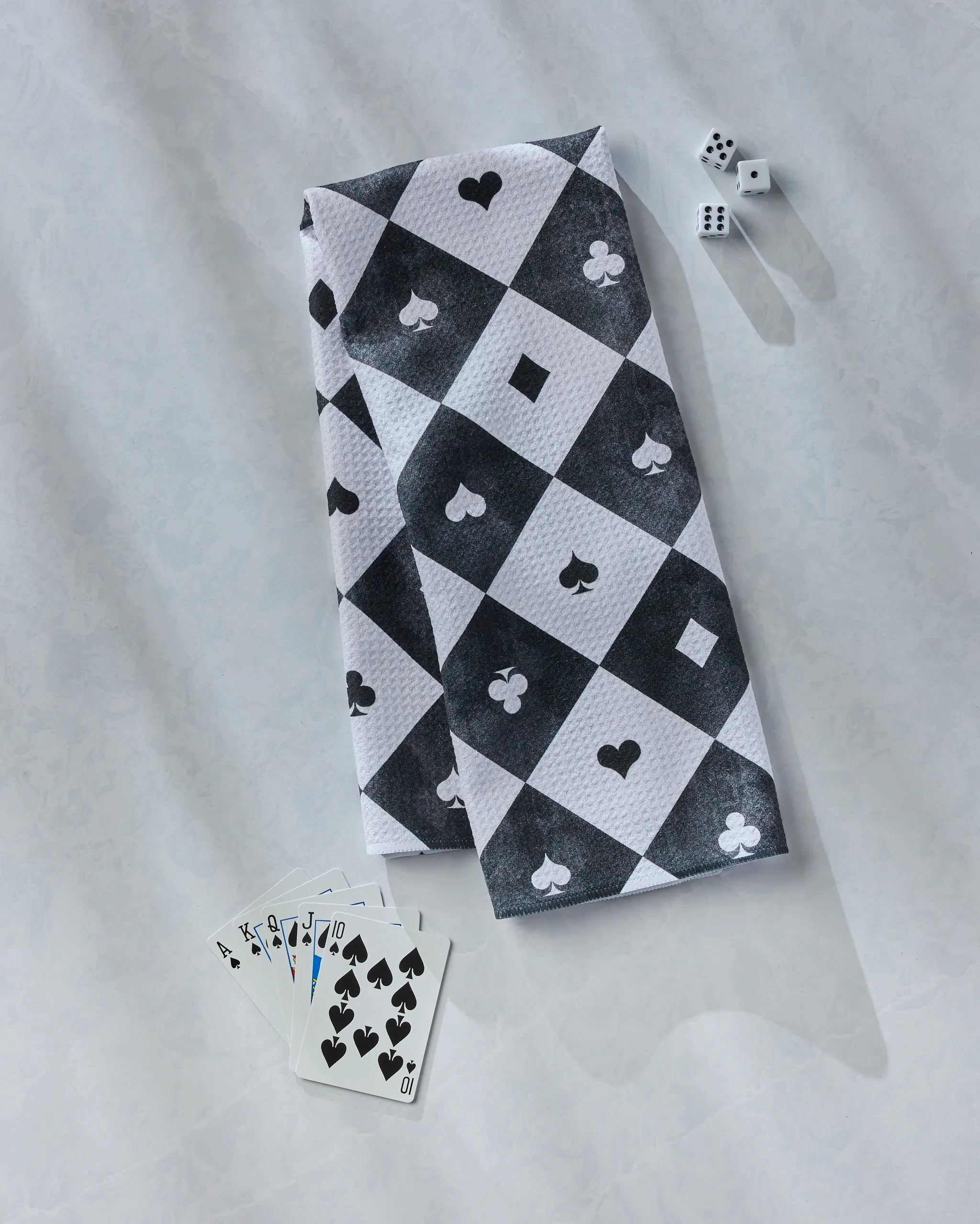 The Geometry "Queen of Everything" kitchen towel, featuring black and white checkers with card suit symbols, is displayed beside three dice and a fanned royal flush in spades.