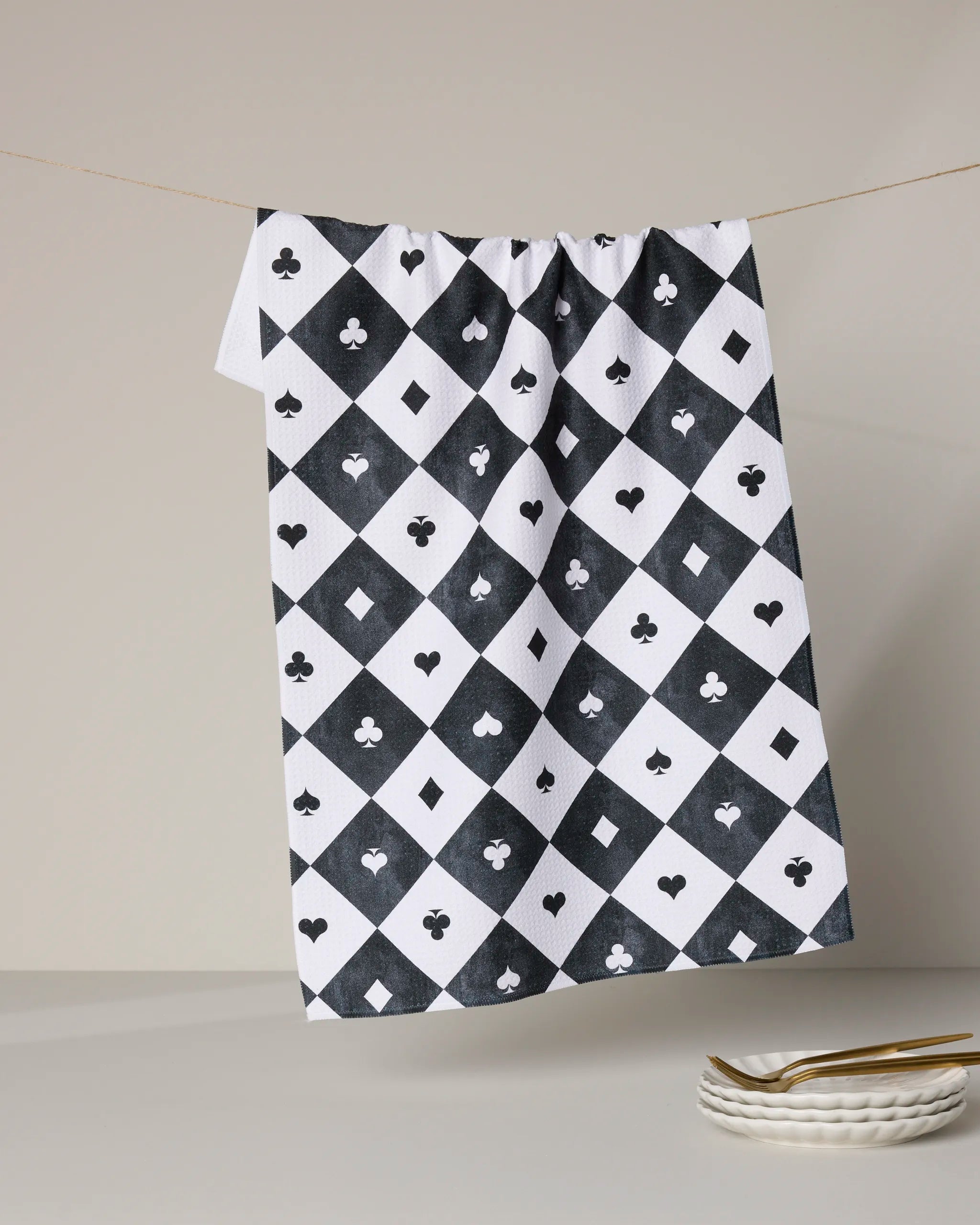 The Queen of Everything kitchen towel by Geometry, made from recycled materials, features black and white diamond patterns with playing card suits and hangs above stacked plates and gold utensils on a white surface.