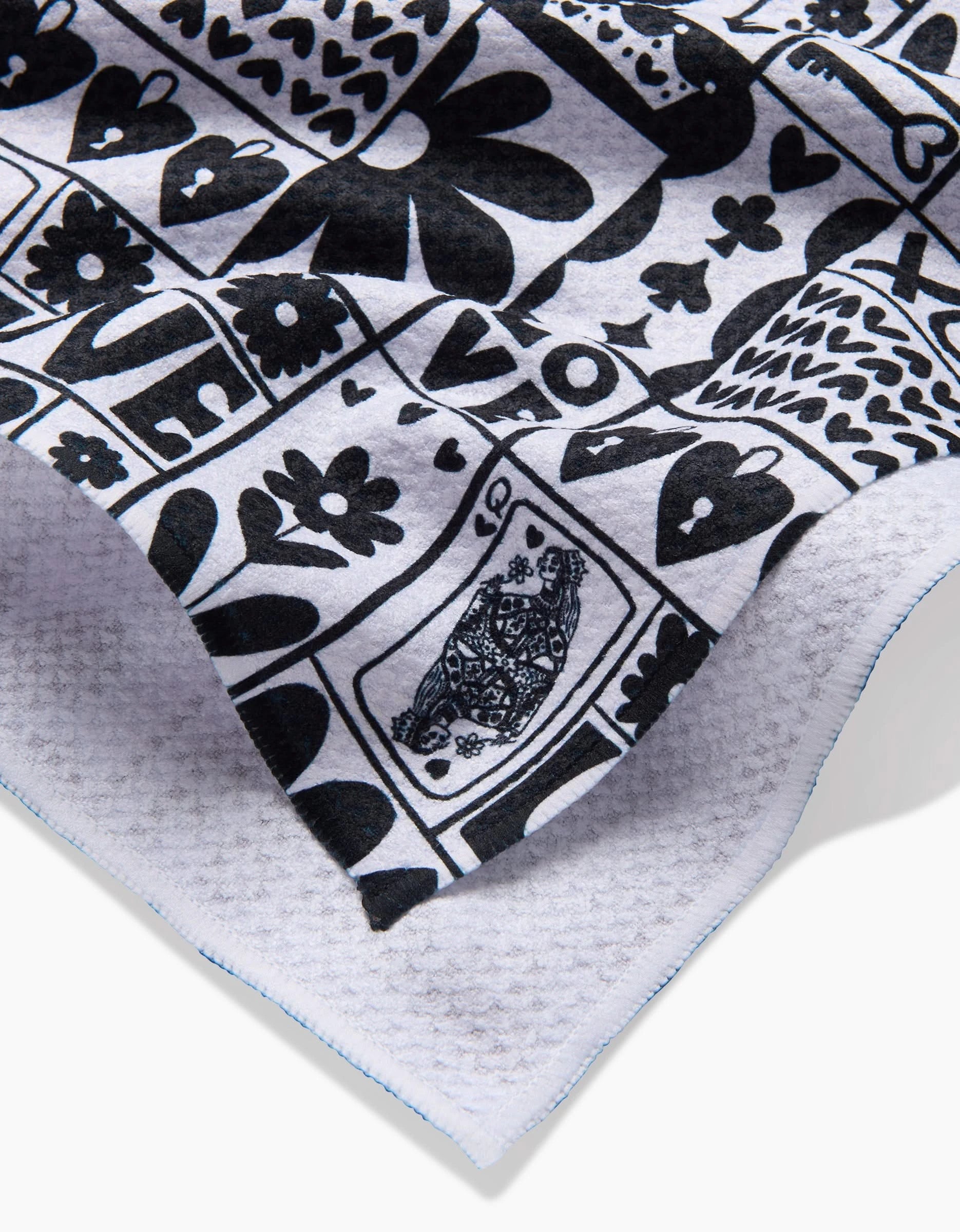 A close-up of the Queen of Hearts tea towel by Kayla Ann shows textured white fabric with black hearts, flowers, a bird, LOVE, and whimsical designs. Blue stitching outlines the edge. Made from recycled materials.