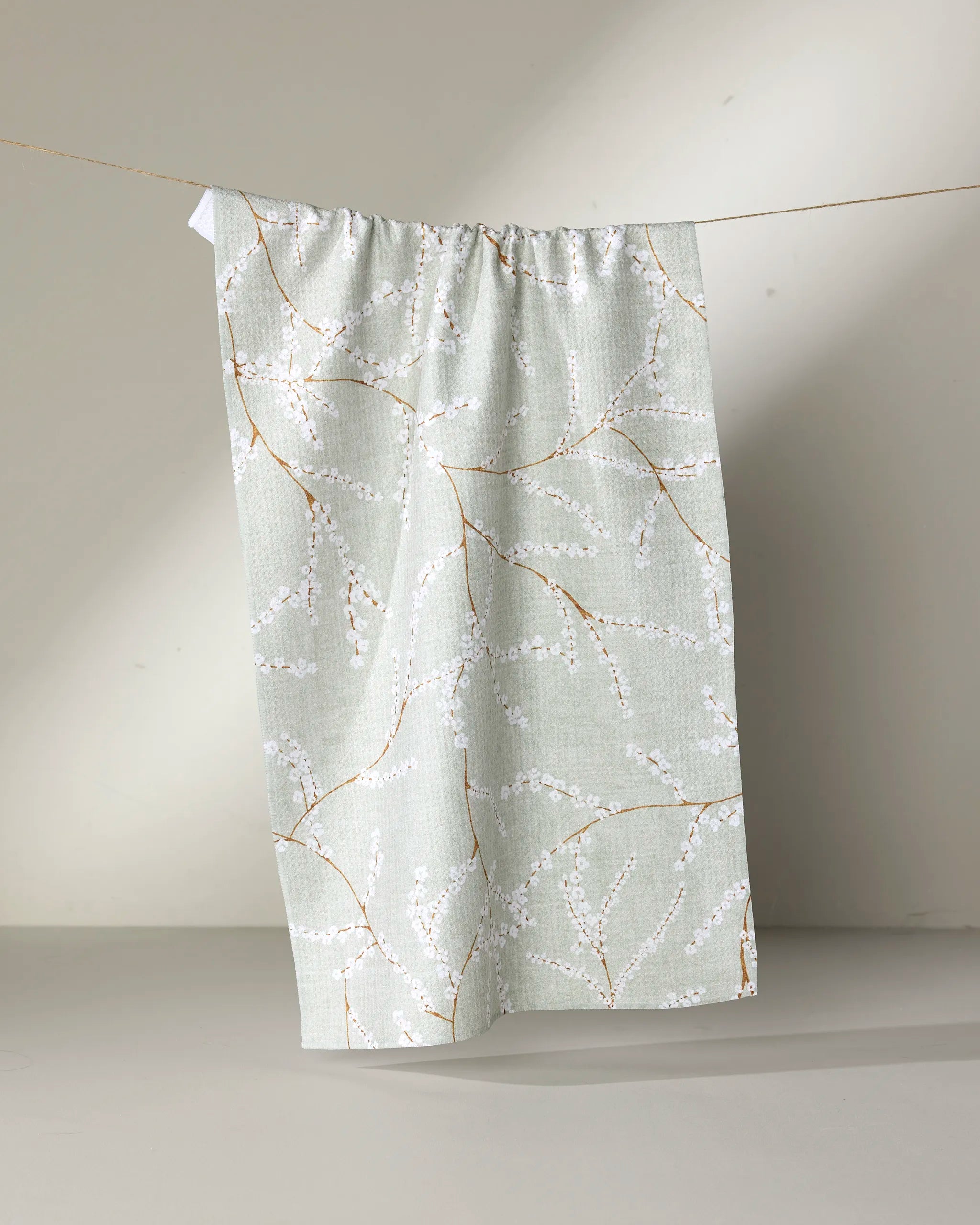 The Quiet Flowers kitchen towel by Pascale Dilger, featuring a pale green shade and white blossom branch pattern, hangs on a string against a neutral background.