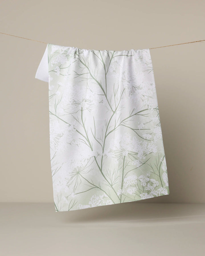 The Quiet Garden tea towel by Geometry, featuring light green and white botanical designs and made from recycled materials, hangs on a string against a plain beige background.