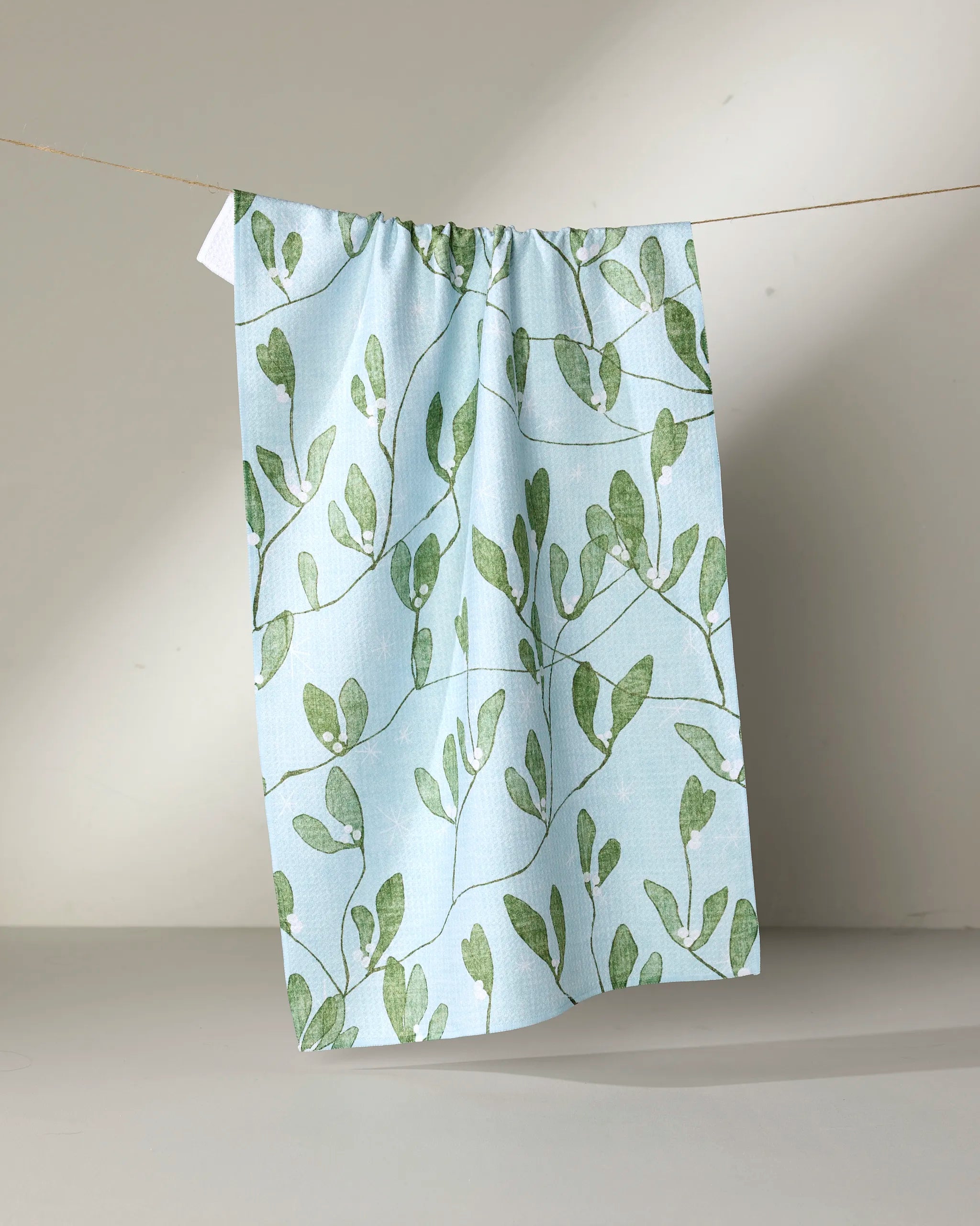 The Quiet Morning tea towel by Pascale Dilger, featuring a green leafy vine pattern on light blue fabric, hangs on a string against a plain beige background.