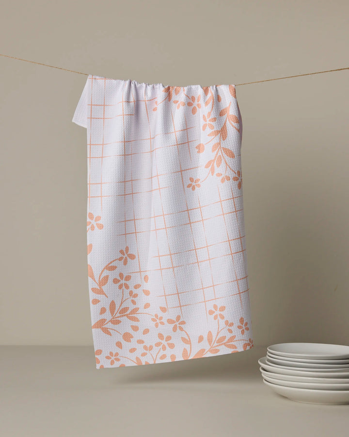 The Quiet Picnic kitchen towel by Geometry, featuring a peach checkered pattern and floral design made from recycled materials, hangs on a string above white plates against a beige background.