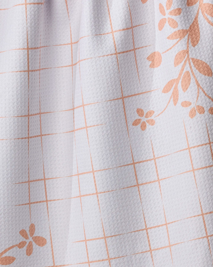 Close-up of the Geometry "Quiet Picnic" kitchen towel, featuring a peach grid pattern with diagonal floral designs of small flowers and leaves on textured fabric.
