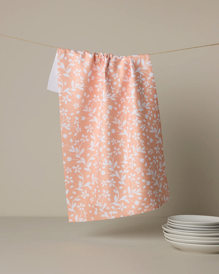 A peach-colored Quiet Radiance tea towel by Geometry, featuring a white floral Geoweave pattern, hangs above a light gray surface beside a small stack of white plates.