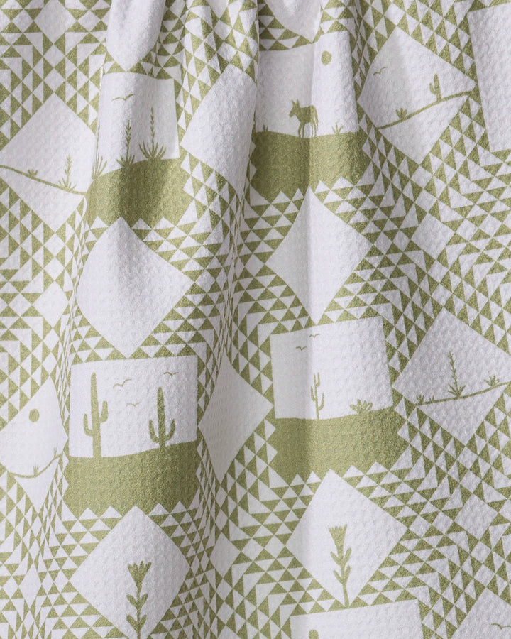 The Elana Gabrielle "Quilt Landscape" tea towel features textured white fabric with green geometric shapes, cacti, desert plants, mountains, and animal silhouettes for a desert-inspired look. Slightly gathered and highly absorbent—ideal for drying dishes.