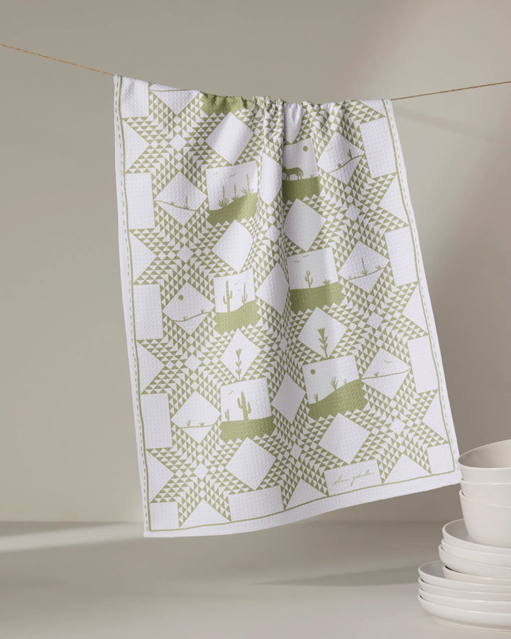 The Elana Gabrielle Quilt Landscape kitchen towel, featuring eco-friendly green and white geometric and desert-inspired patterns, hangs above stacked white dishes set against a neutral background.