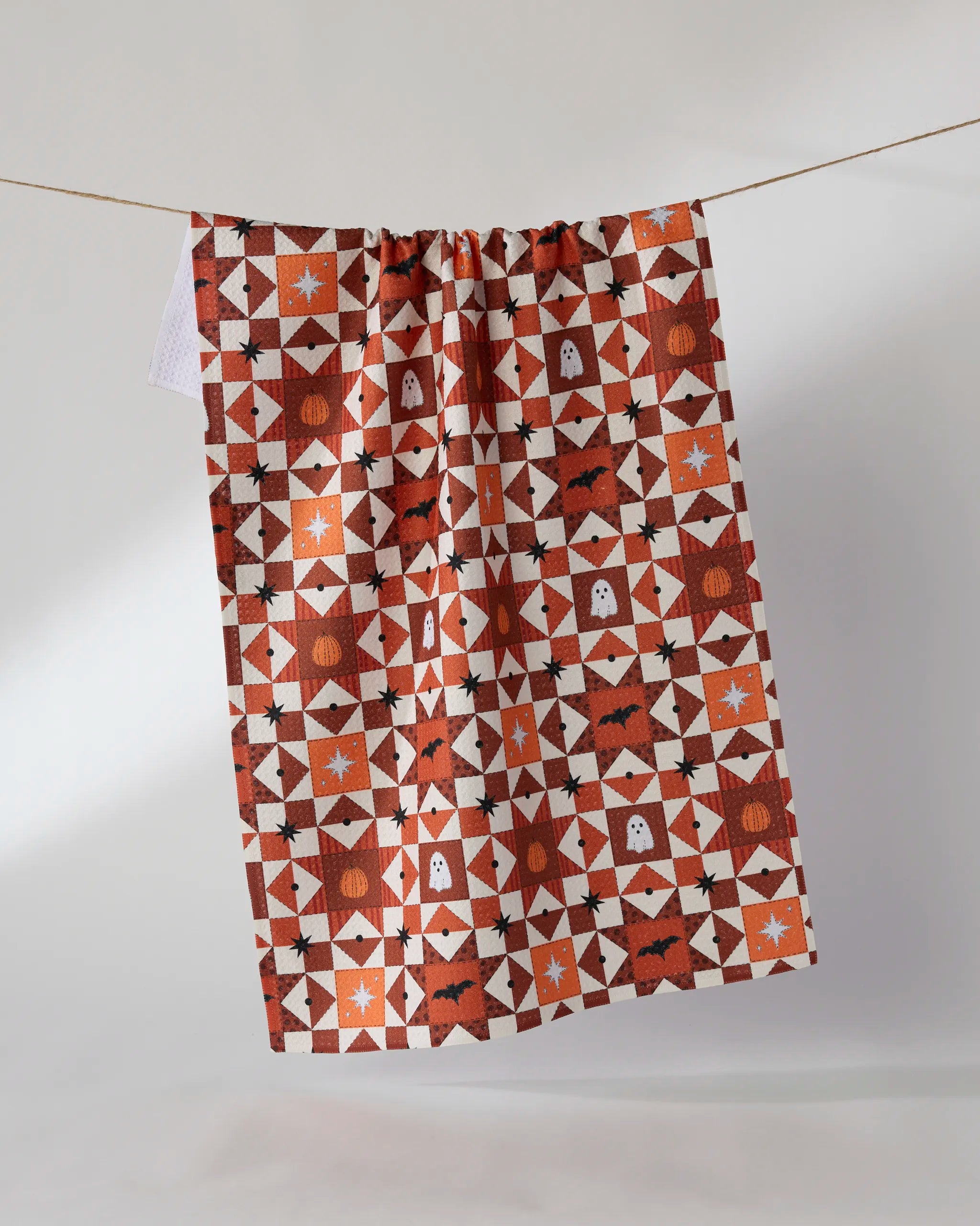 The Geometry "Quilt Work" kitchen towel, featuring a geometric patchwork design in orange, brown, and white with Halloween motifs like bats, ghosts, stars, and pumpkins, hangs on a line against a plain background.