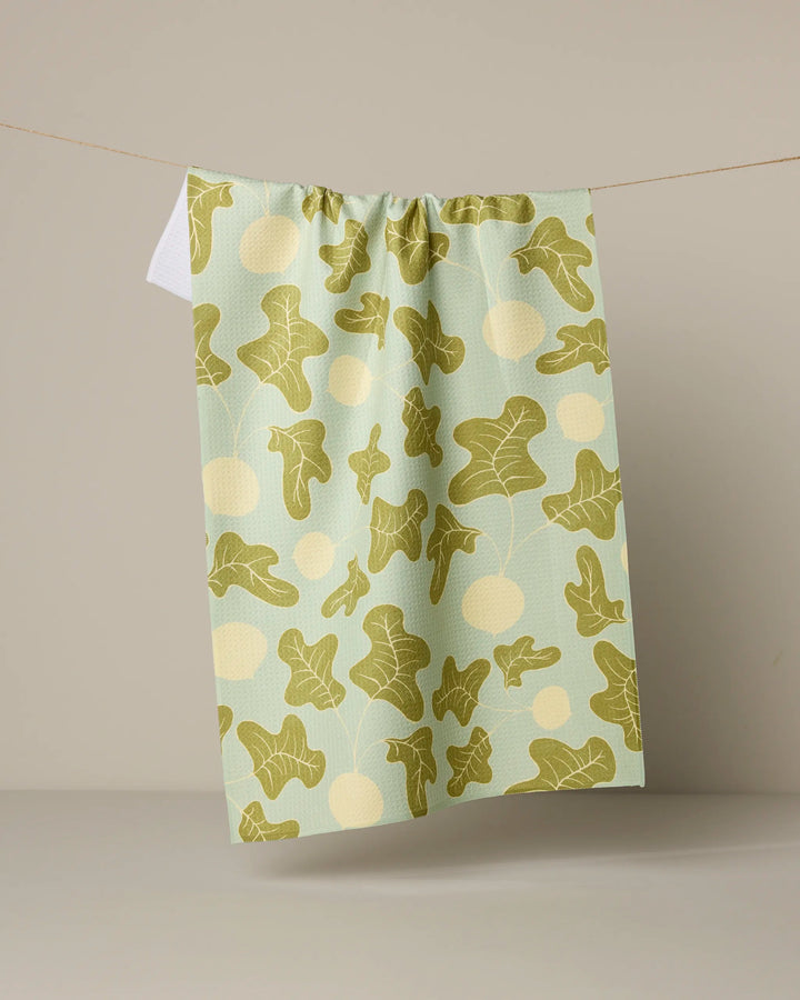 The Geometry Radish Pop kitchen tea towel, made from recycled materials, displays a vibrant radish and polka dot pattern in green and yellow, hanging against a beige background.