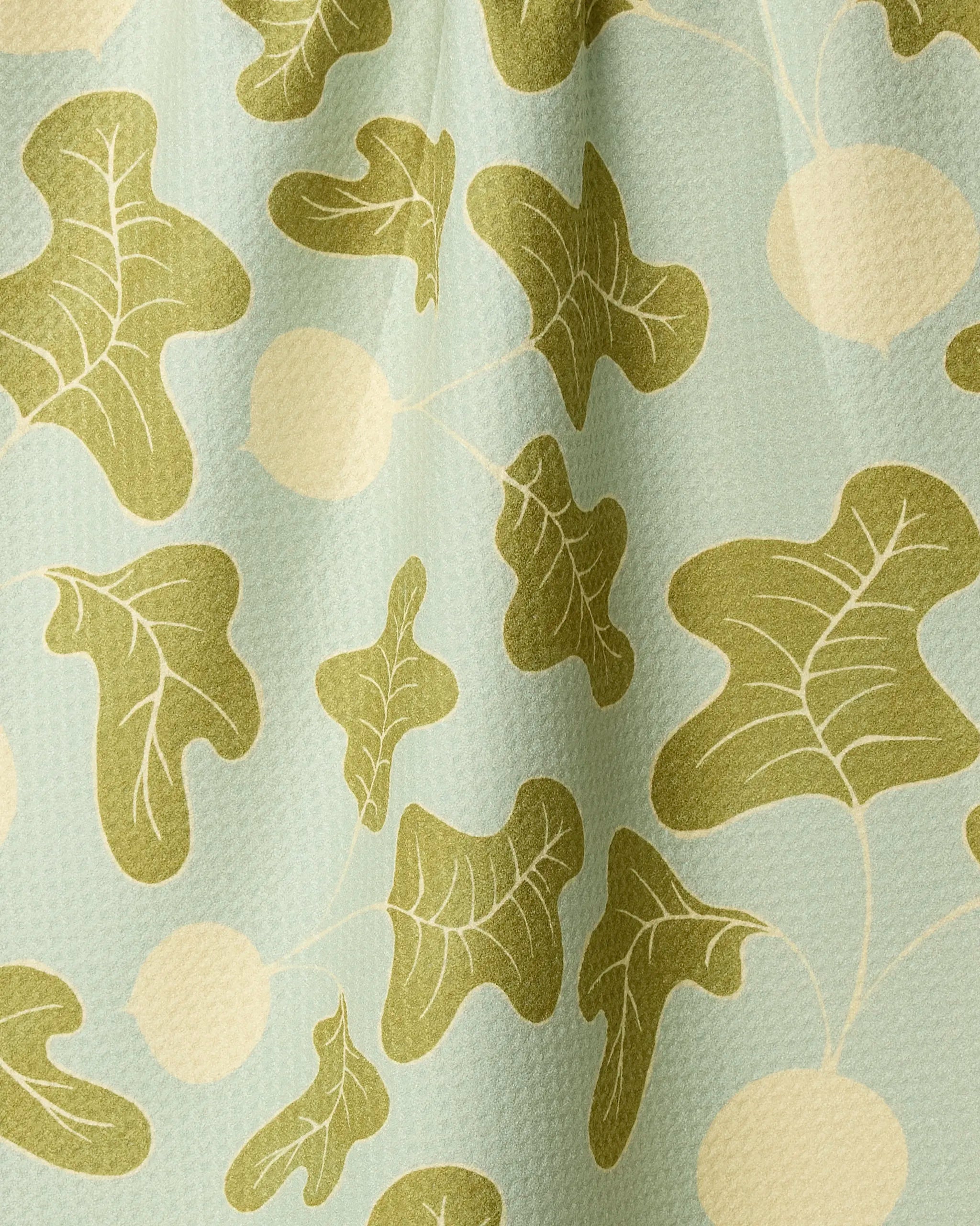 The Geometry Radish Pop tea towel features a patterned fabric with olive green leaves and cream circles on a light blue background. Made from recycled materials, it has a subtly textured surface and soft folds.